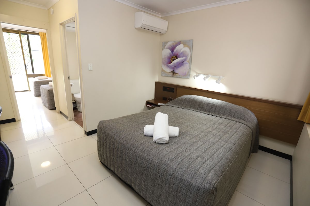 Quarto de hotel com cama, mesa de cabeceira, um quadro e banheiro visível por uma porta aberta.
