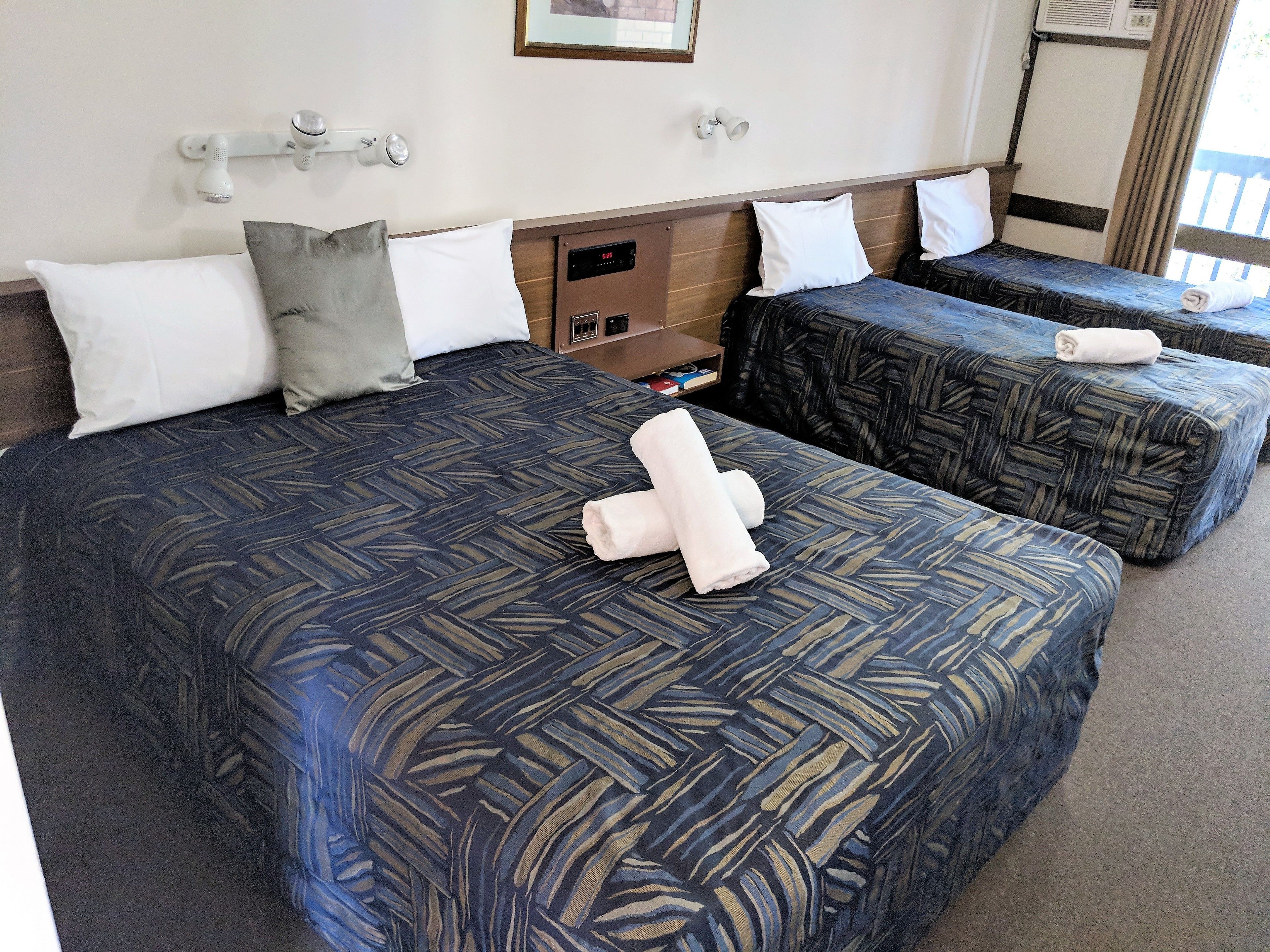 Habitación de hotel con dos camas, cada una con una colcha y almohadas estampadas. Se observa una pequeña estantería con artículos de aseo y un reloj en la pared.