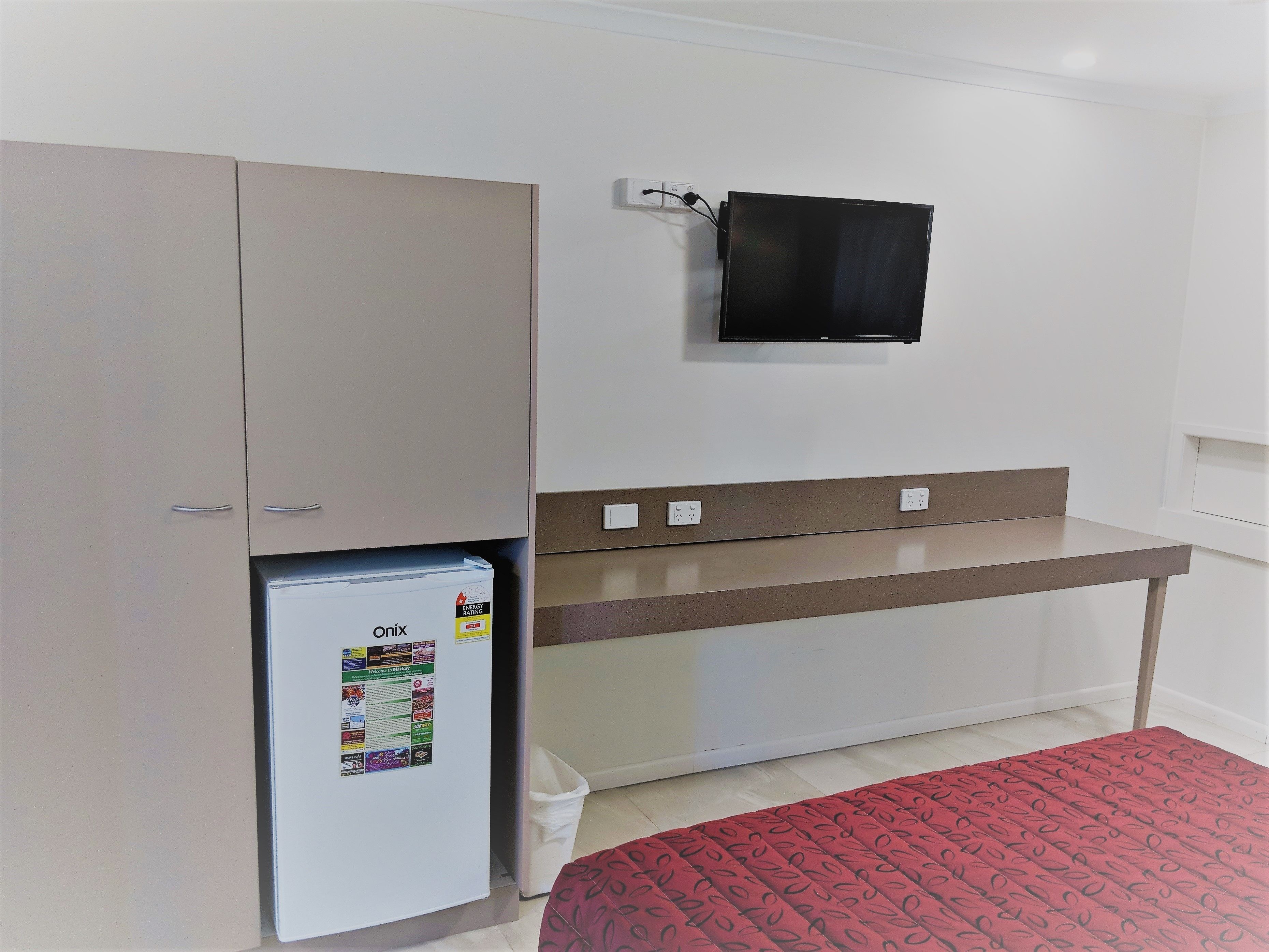 A compact room with a kitchenette, a small refrigerator, a flat-screen TV mounted on the wall, and a bed with a patterned cover.