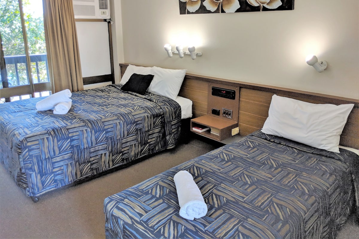 Quarto de hotel com duas camas, uma mesa de cabeceira, ar condicionado fixado na parede e uma pintura de flores.