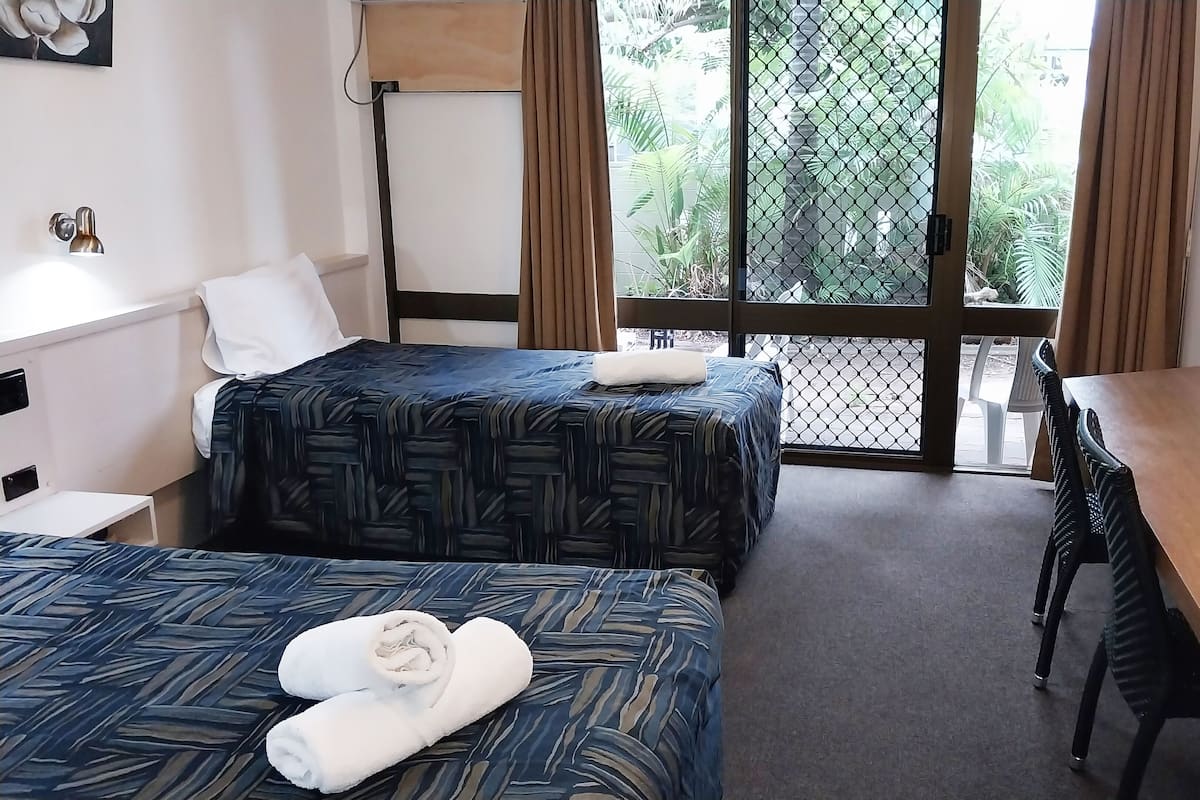 Quarto de hotel com duas camas, uma mesa de trabalho, televisão e varanda com vista para áreas verdes.