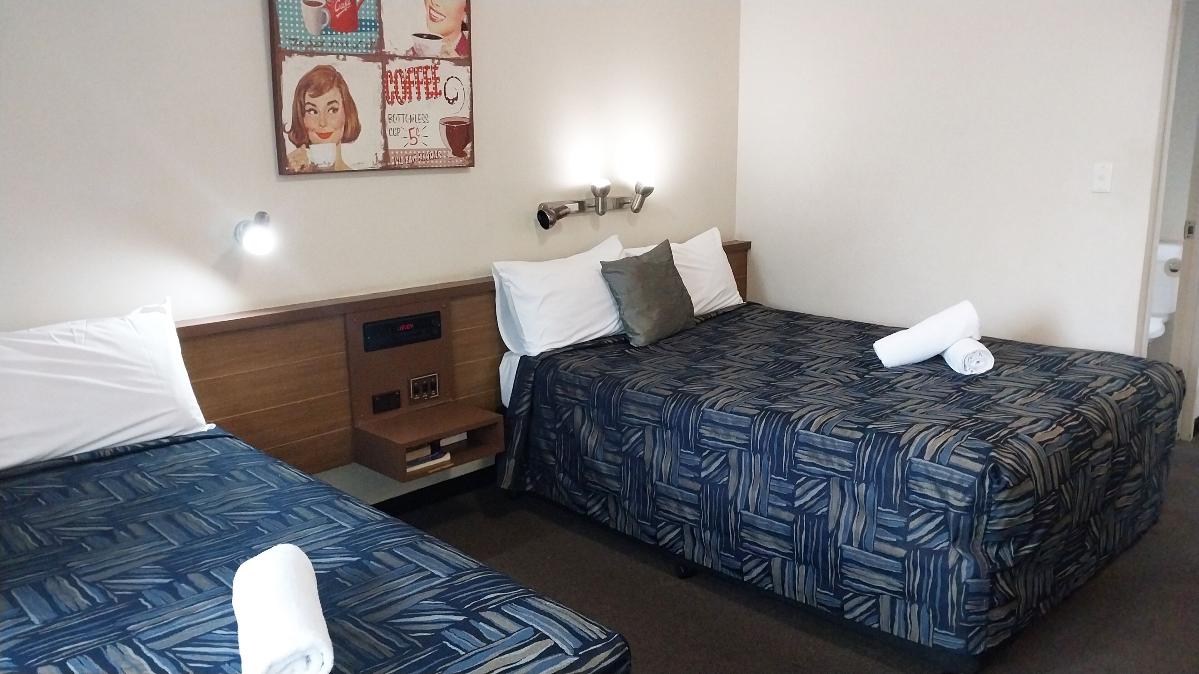 Habitación de hotel con dos camas, cada una con una colcha azul estampada, almohadas blancas y una toalla enrollada. Hay un aplique en la pared sobre las camas y un pequeño estante con un reloj y algunos objetos.