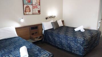 Quarto de hotel com duas camas, cada uma com colcha azul estampada, almofadas brancas e uma toalha enrolada. Há um aplique acima das camas e uma pequena prateleira com um relógio e alguns objetos.