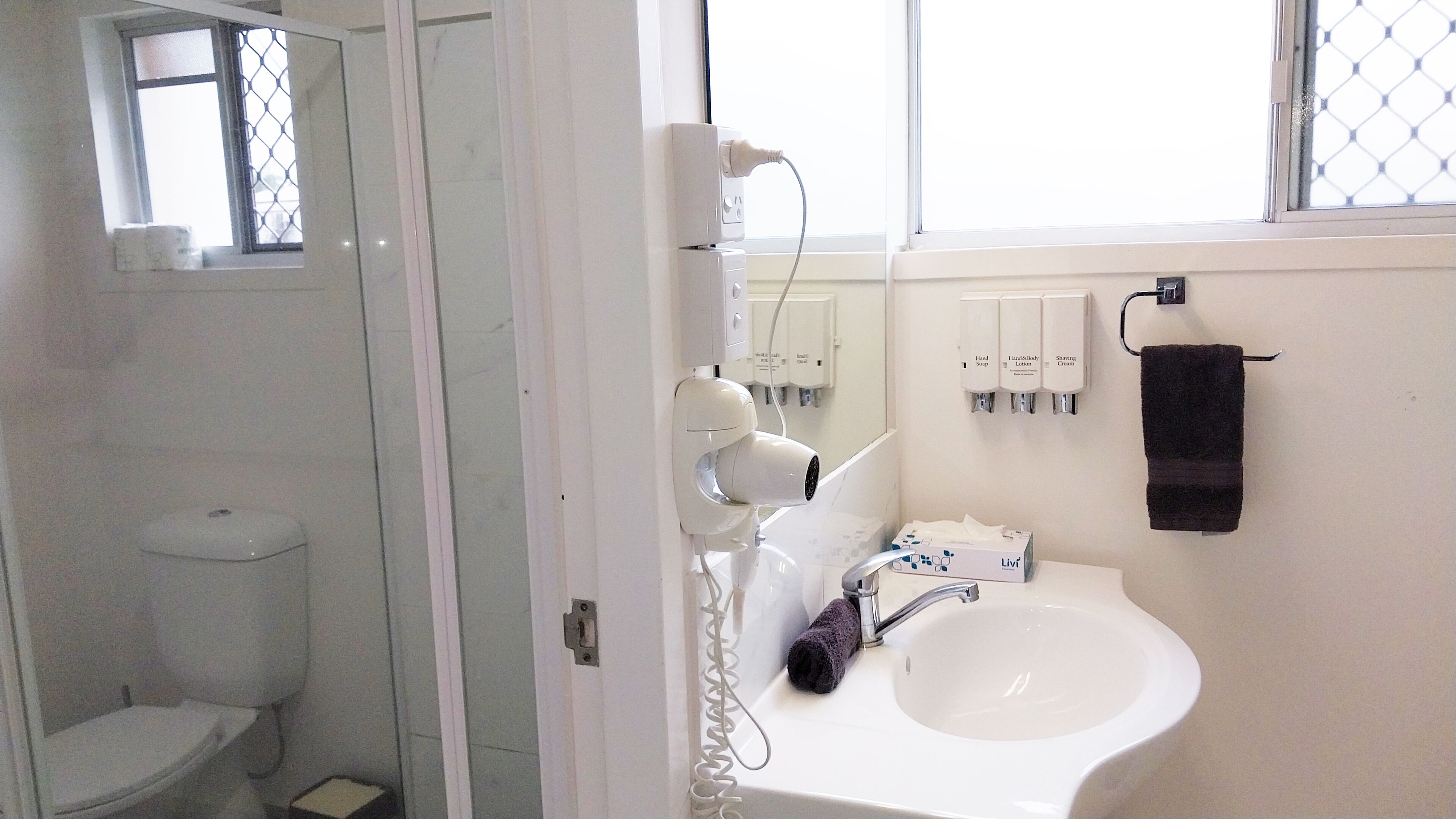 A bathroom with a toilet, a sink, a hairdryer, and hand soap dispensers.
