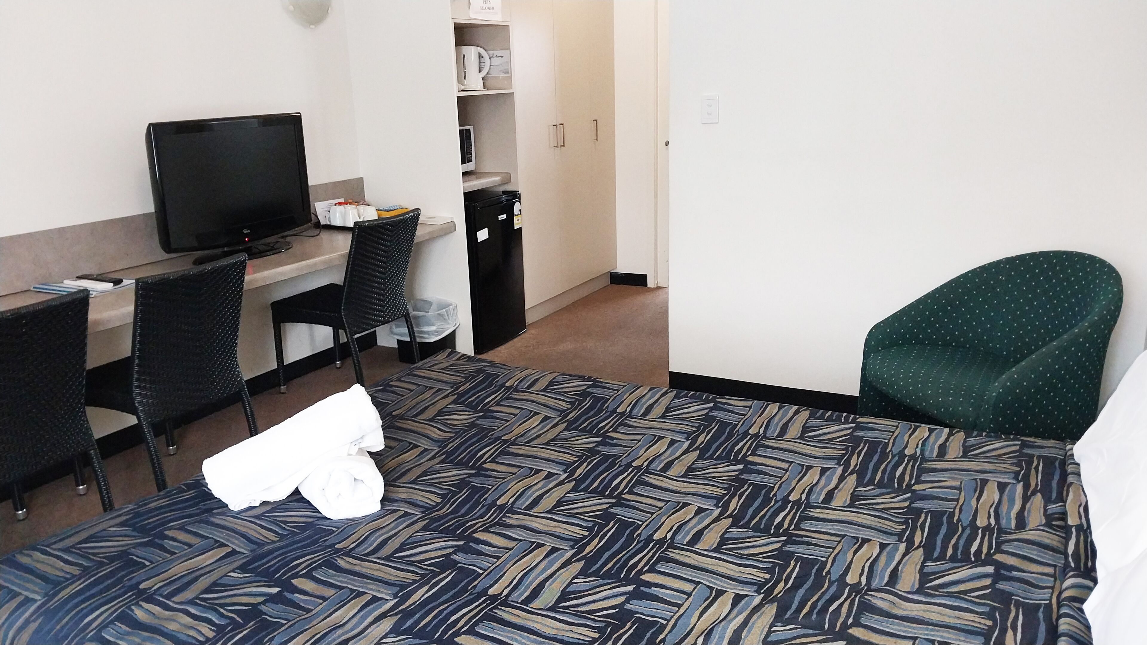 A hotel room with a bed, a desk with a computer, a chair, a TV, and a green armchair.