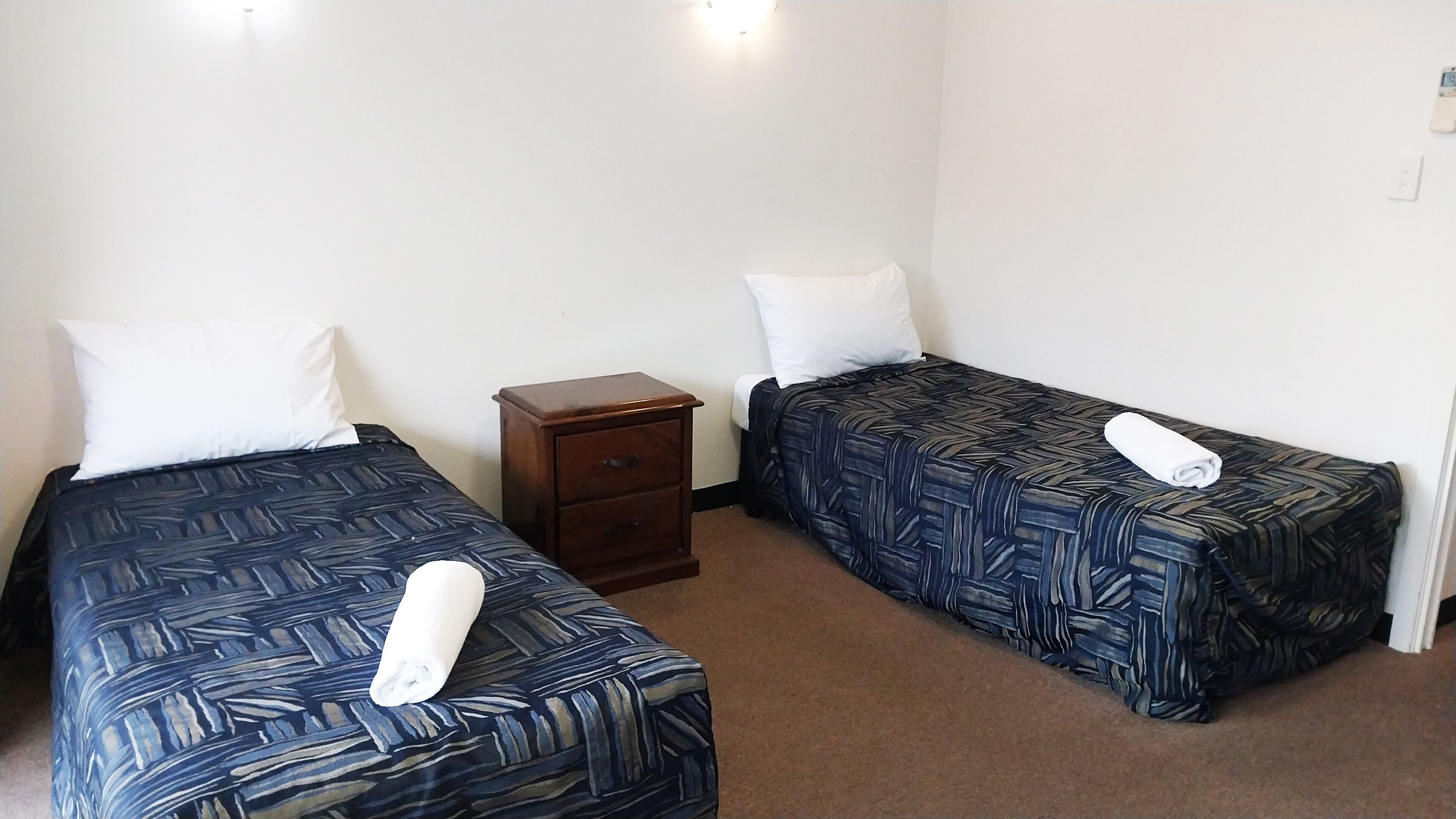 Two single beds with patterned bedding, a small wooden nightstand, and a white wall with a light fixture.