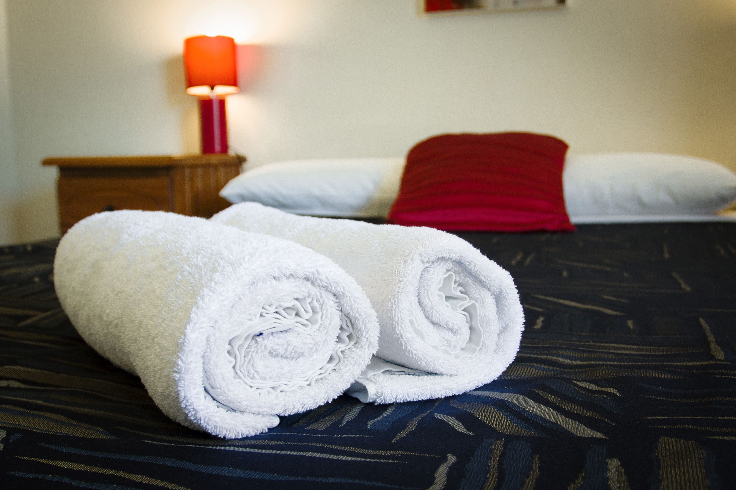 Una cama bien hecha con almohadas blancas y rojas, una toalla blanca enrollada y una lámpara roja sobre una mesita de noche de madera.
