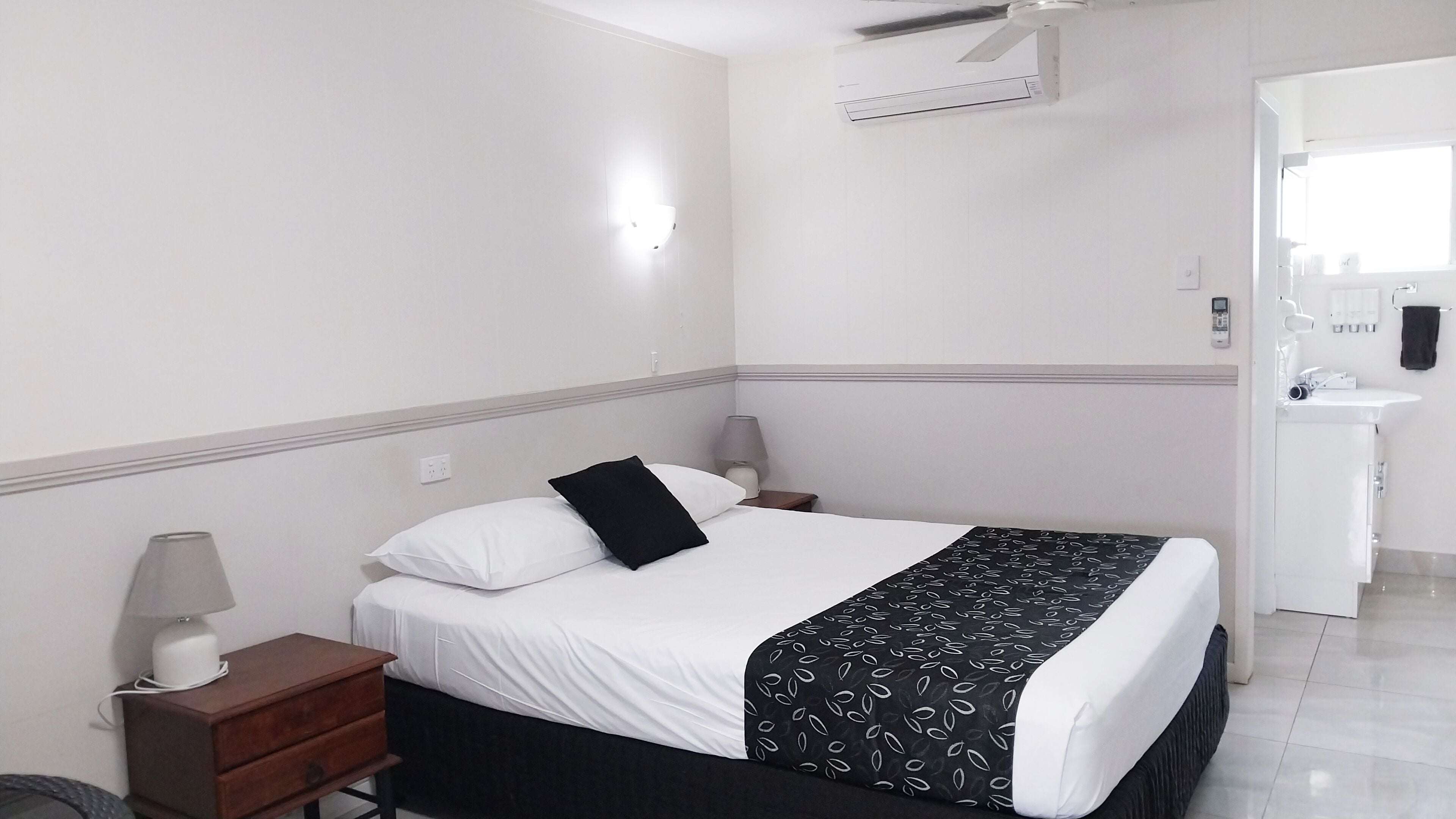 A hotel room with a bed, bedside tables, a lamp, an air conditioning unit, and a bathroom with a sink and mirror.
