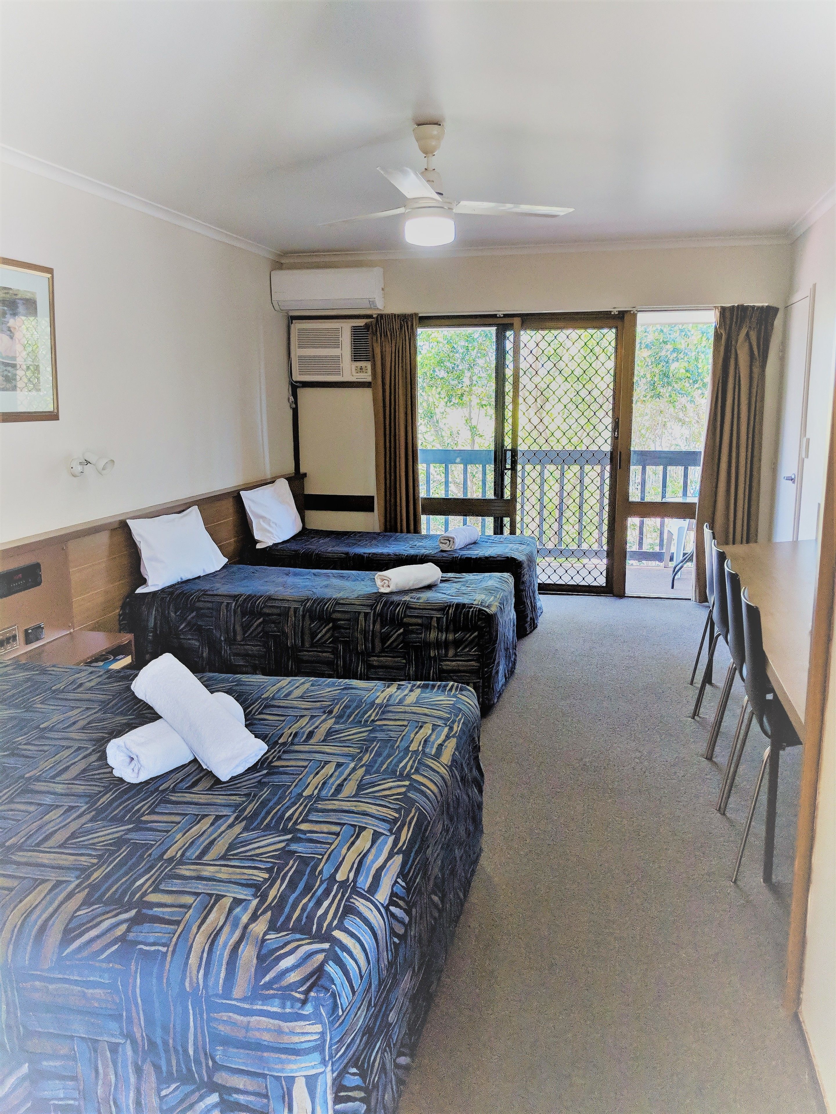 A hotel room with three beds, a ceiling fan, an air conditioning unit, a view of the outside balcony, and a table with chairs.