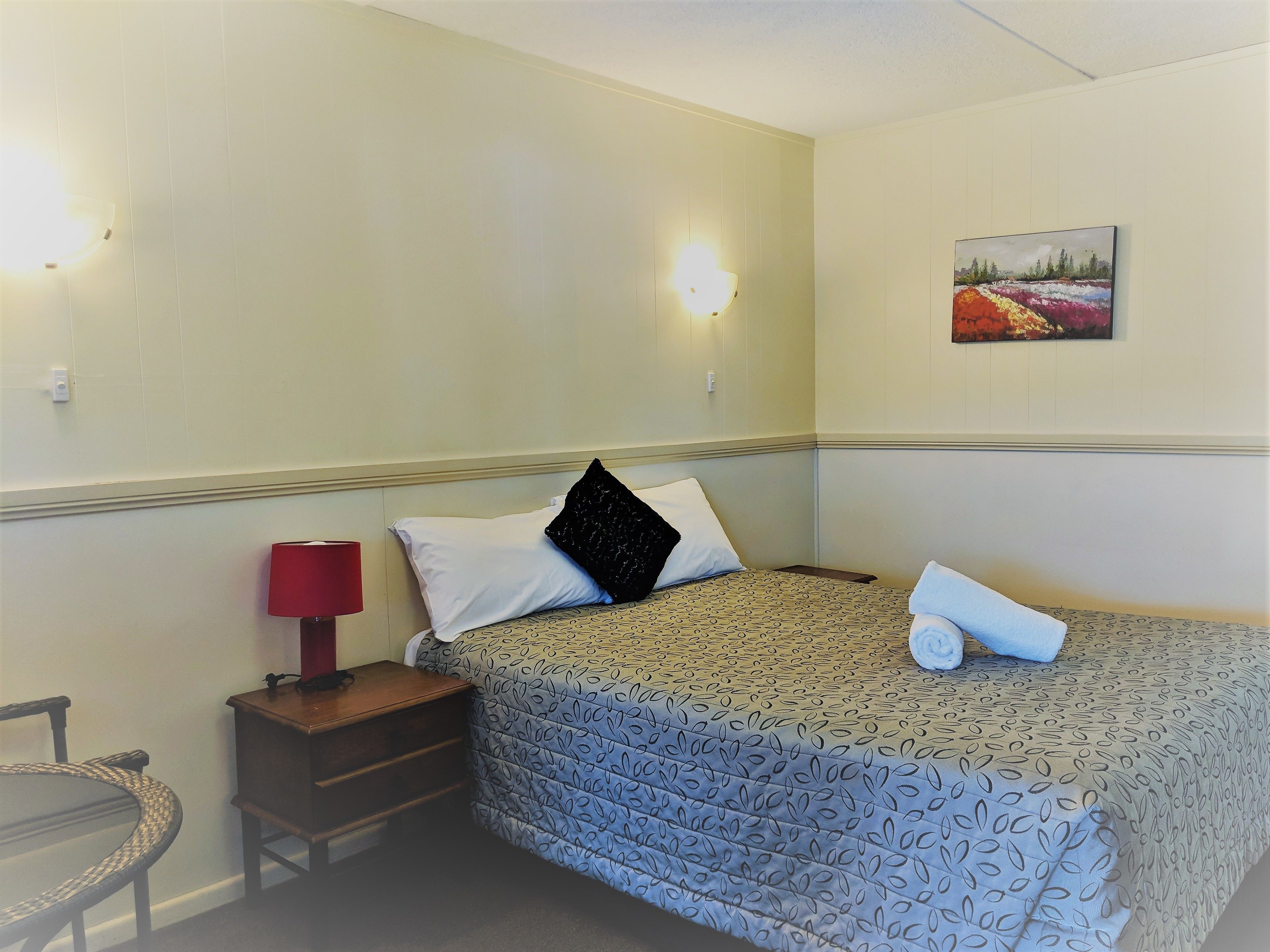 Habitación de hotel con cama, mesita de noche, lámpara roja y un cuadro en la pared.
