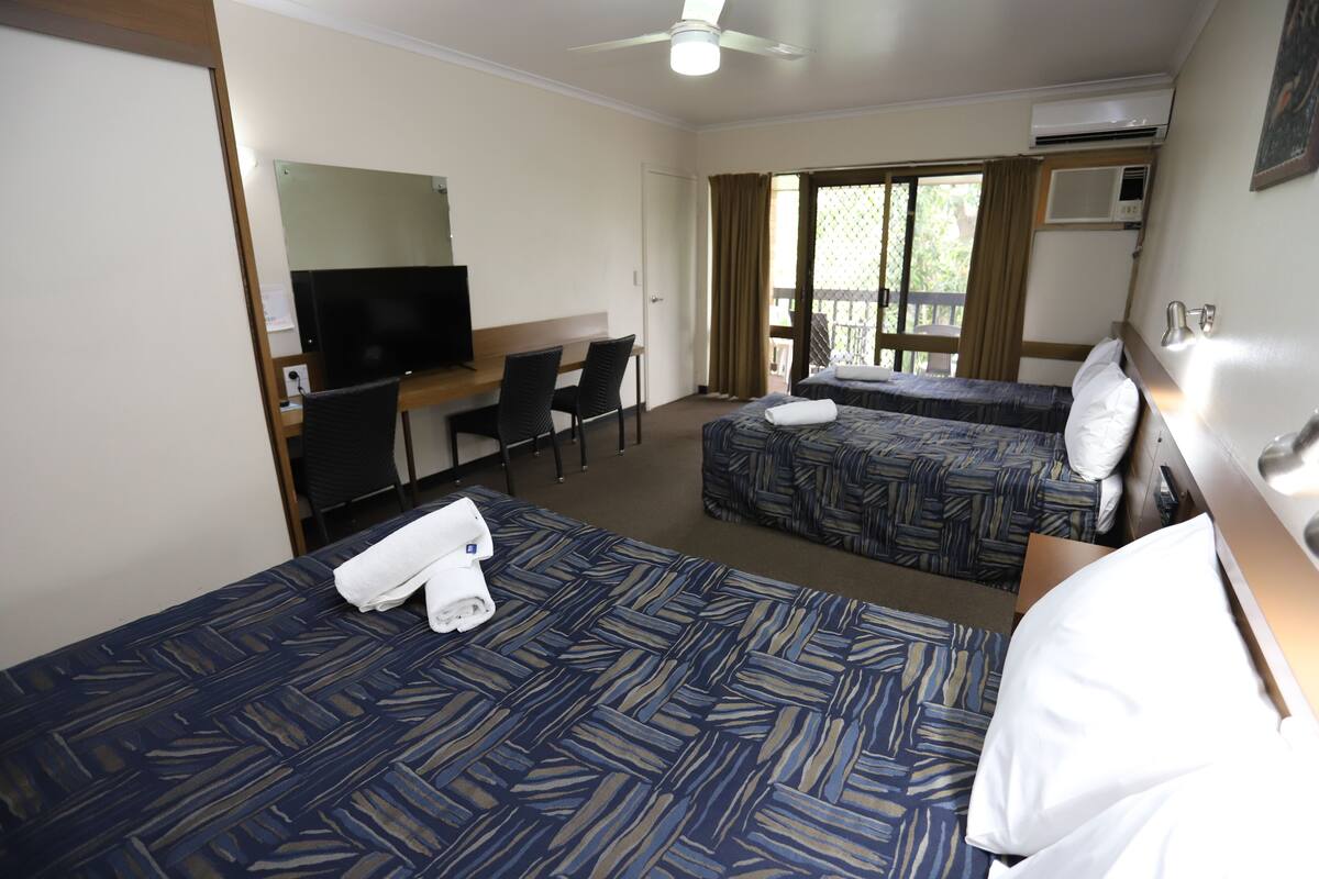 Quarto de hotel com duas camas, uma escrivaninha com cadeira, televisão e varanda com cortinas.