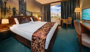Una habitación de hotel con una cama grande, dos mesitas de noche con lámparas, un escritorio con una silla y un ventanal con vistas a edificios.