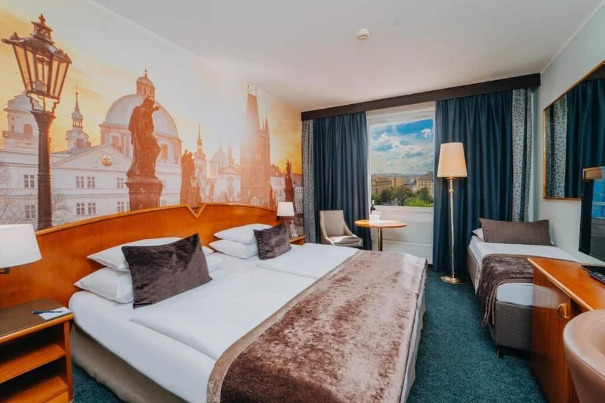 Habitación de hotel con una cama grande, un escritorio, una silla y vistas a una ciudad a través del ventanal.