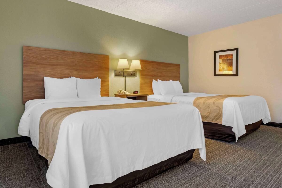 A hotel room with two beds, a wooden headboard, a nightstand with a lamp, and a framed picture on the wall.