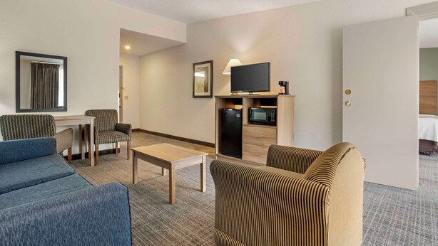 A hotel room with a blue sofa, striped armchair, a small table, a TV on a cabinet, a microwave, and a door leading to a bedroom. - Quality Inn & Suites NRG Park - Medical Center (Houston)