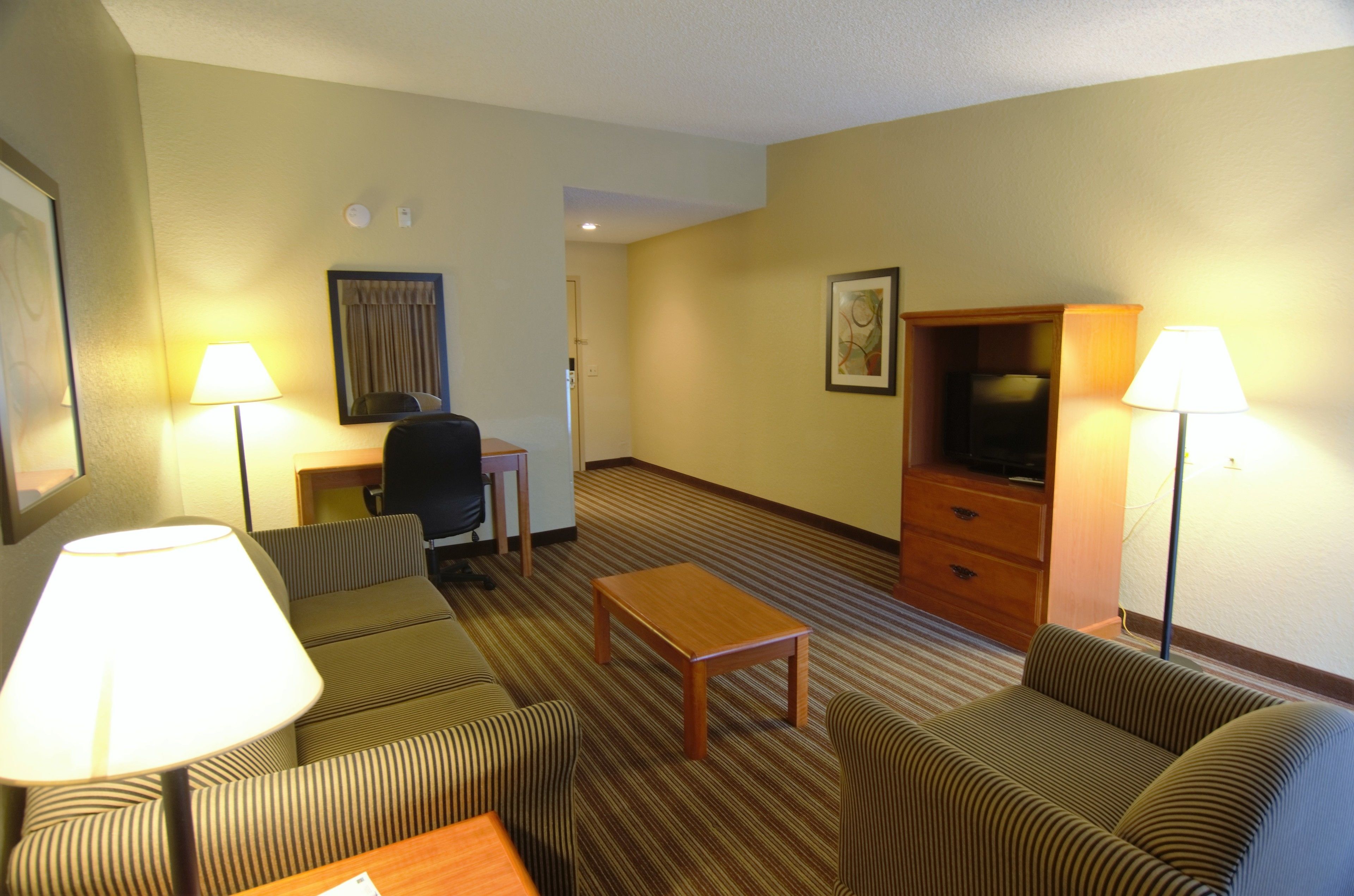 A hotel room with striped sofas, a wooden coffee table, a desk with a chair, a television, and two lamps.