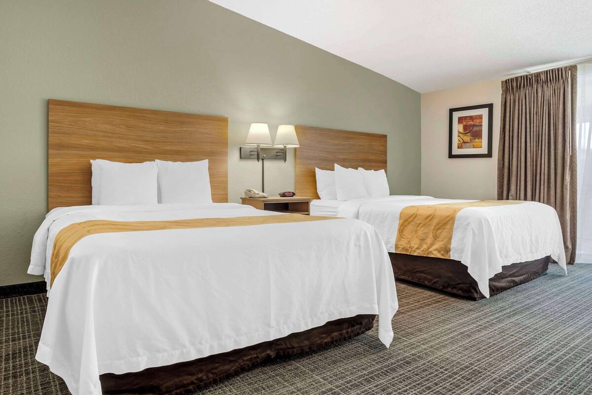 A hotel room with two beds, a headboard, a nightstand with a lamp, a framed picture on the wall, and a window with curtains.