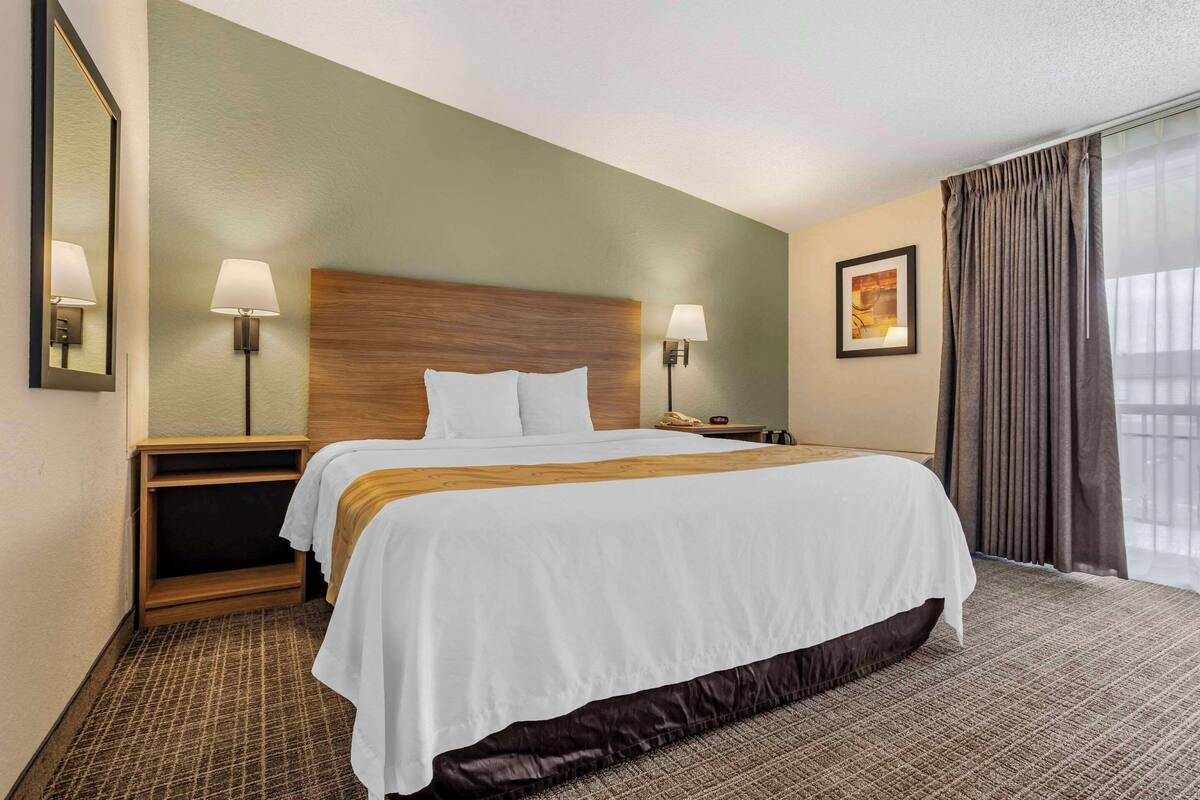 A hotel room with a large bed, two bedside tables, a nightstand, a mirror, and a window with curtains.