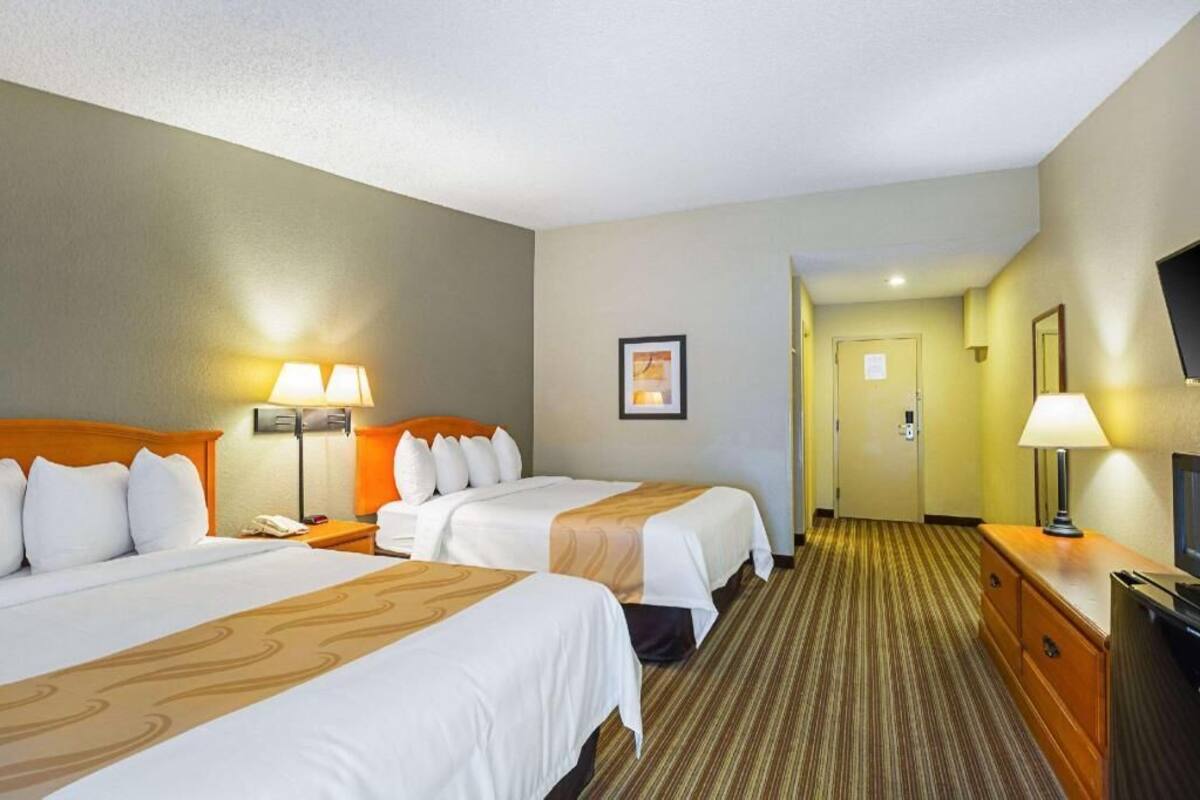 A hotel room with two beds, a television, a desk, and a door leading to a hallway.