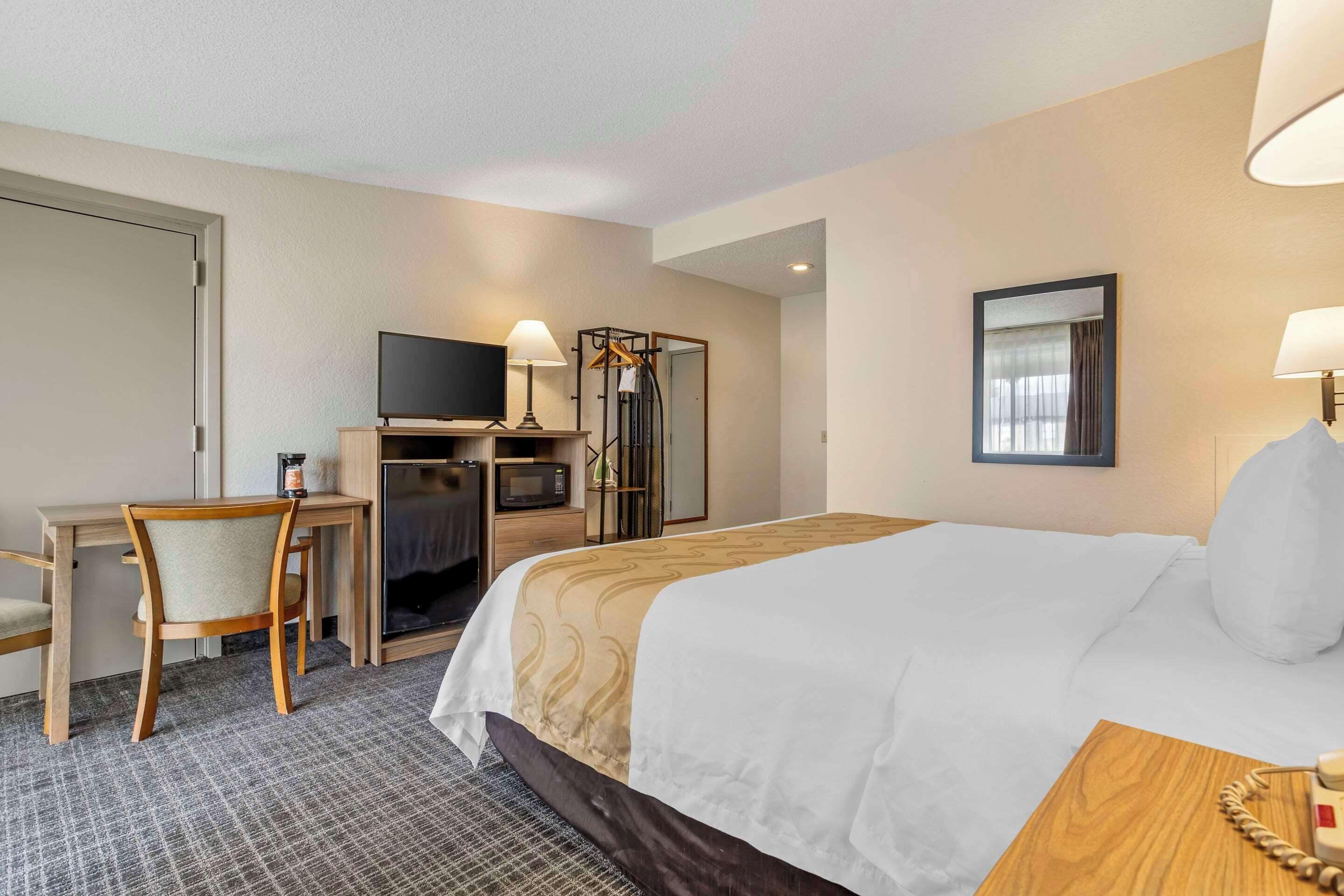 A hotel room with a bed, a desk with a chair, a television, a mirror, and a lamp.