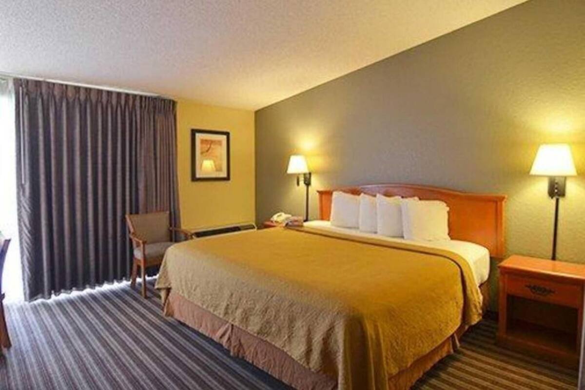 A hotel room with a bed, two bedside lamps, a chair, a desk, and a window with curtains.