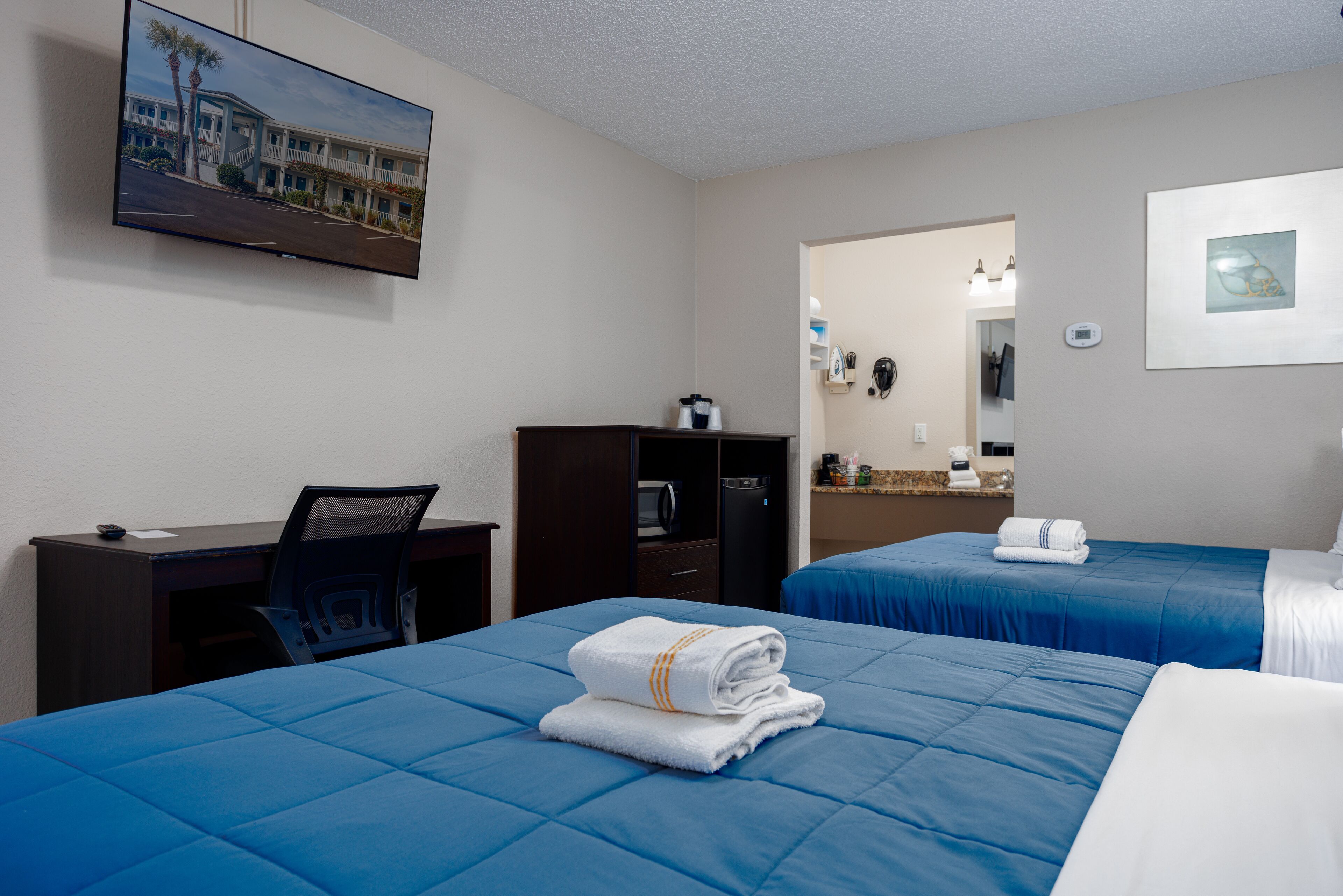 A hotel room with two beds, a desk, a chair, a television, a wardrobe, and a bathroom visible through an open door.