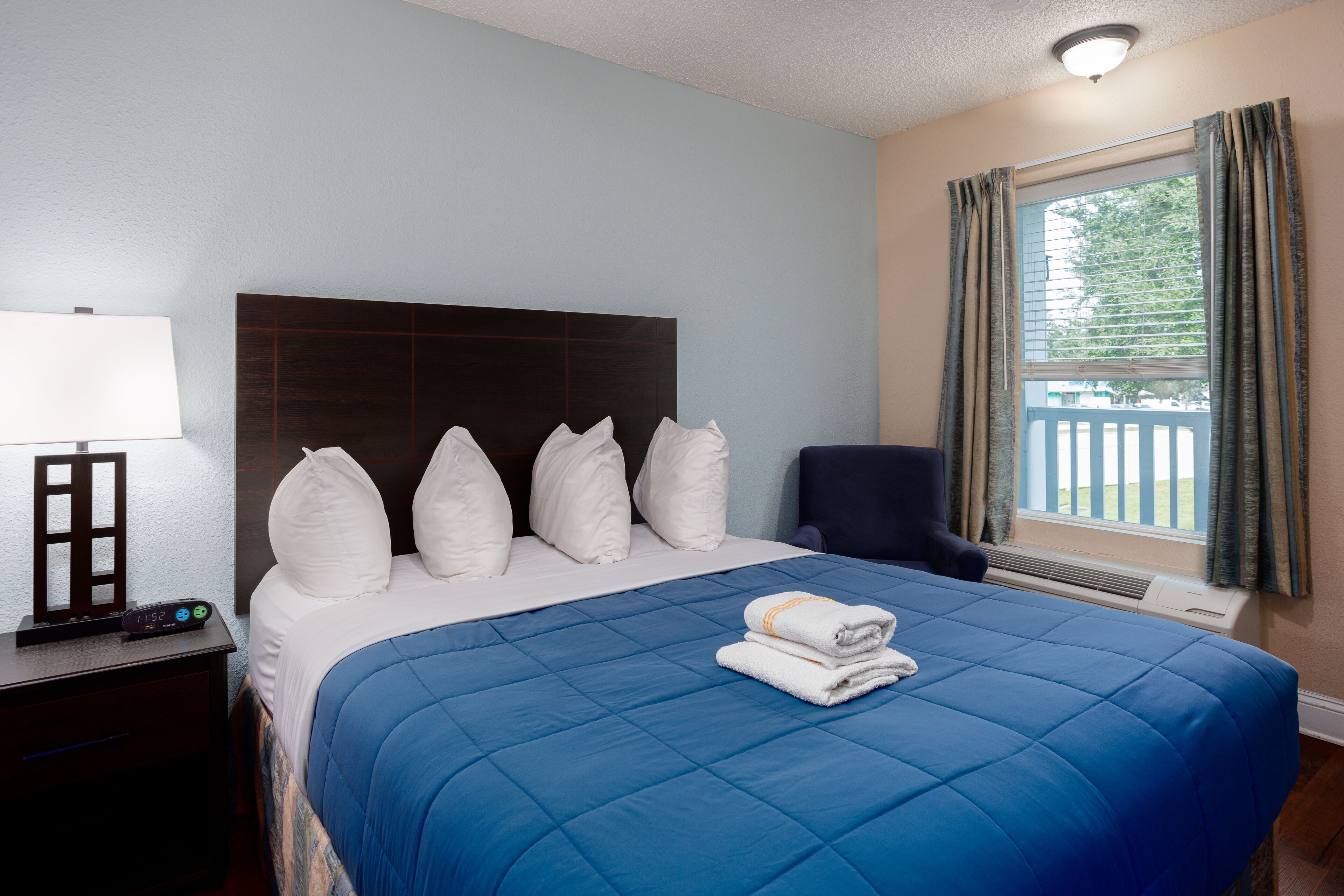 A hotel room with a large bed, a nightstand with a lamp, a chair, and a window with curtains.