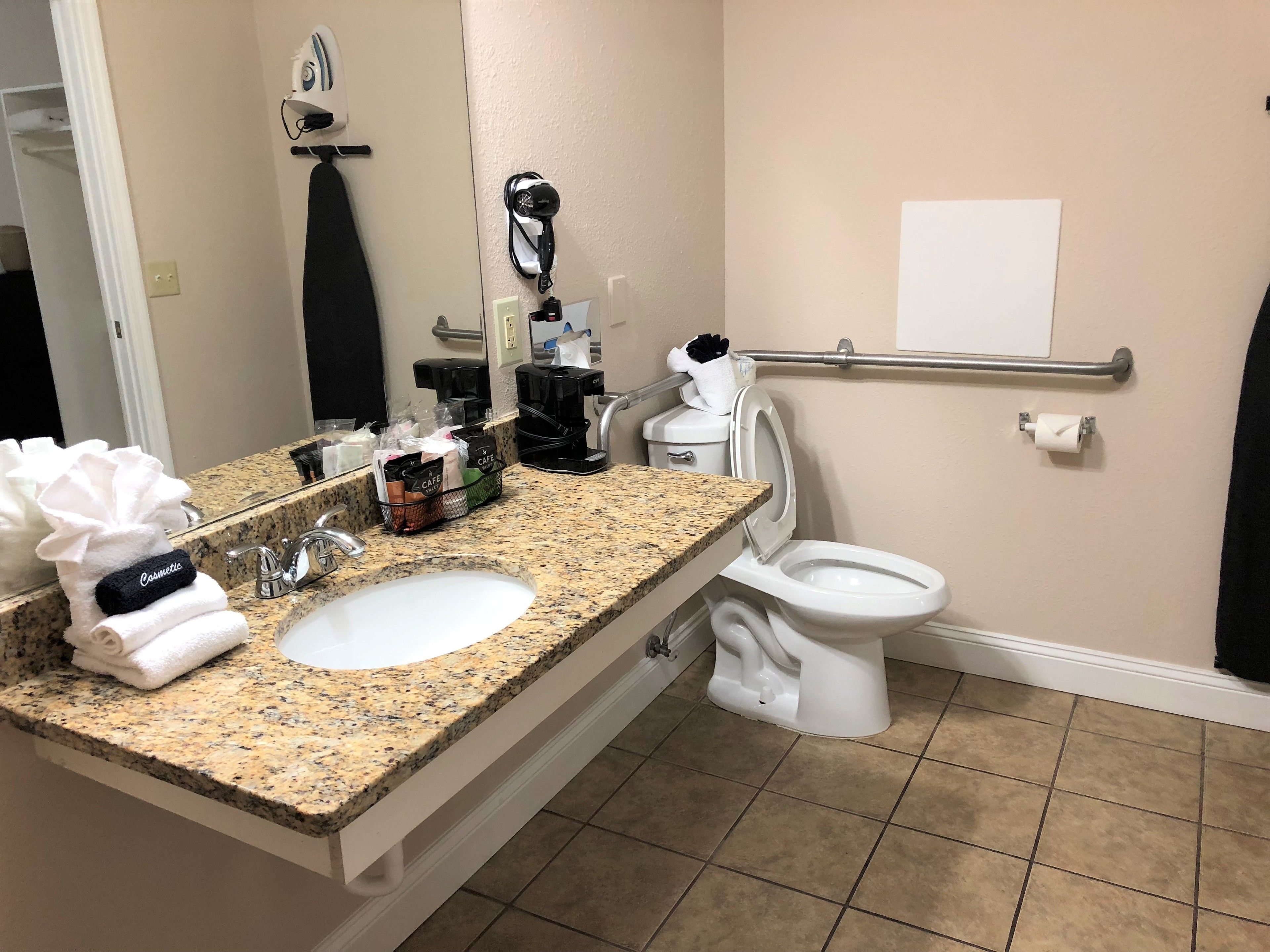 A bathroom with a granite countertop sink, a mirror, a towel rack, a hair dryer, a toilet, and a handrail.