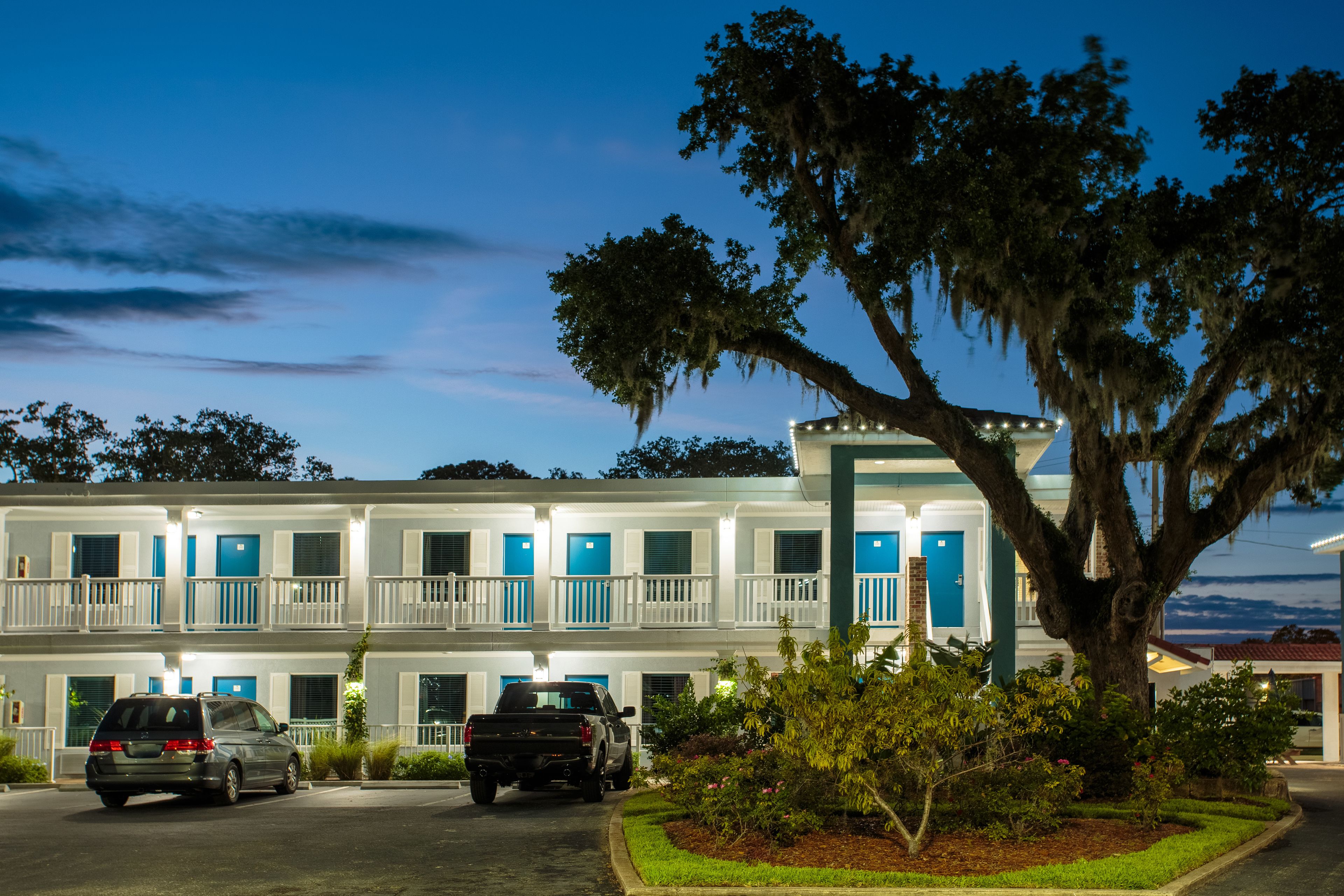 A motel with a two-story structure, multiple rooms with blue doors, a parking area, and a large tree in the foreground.