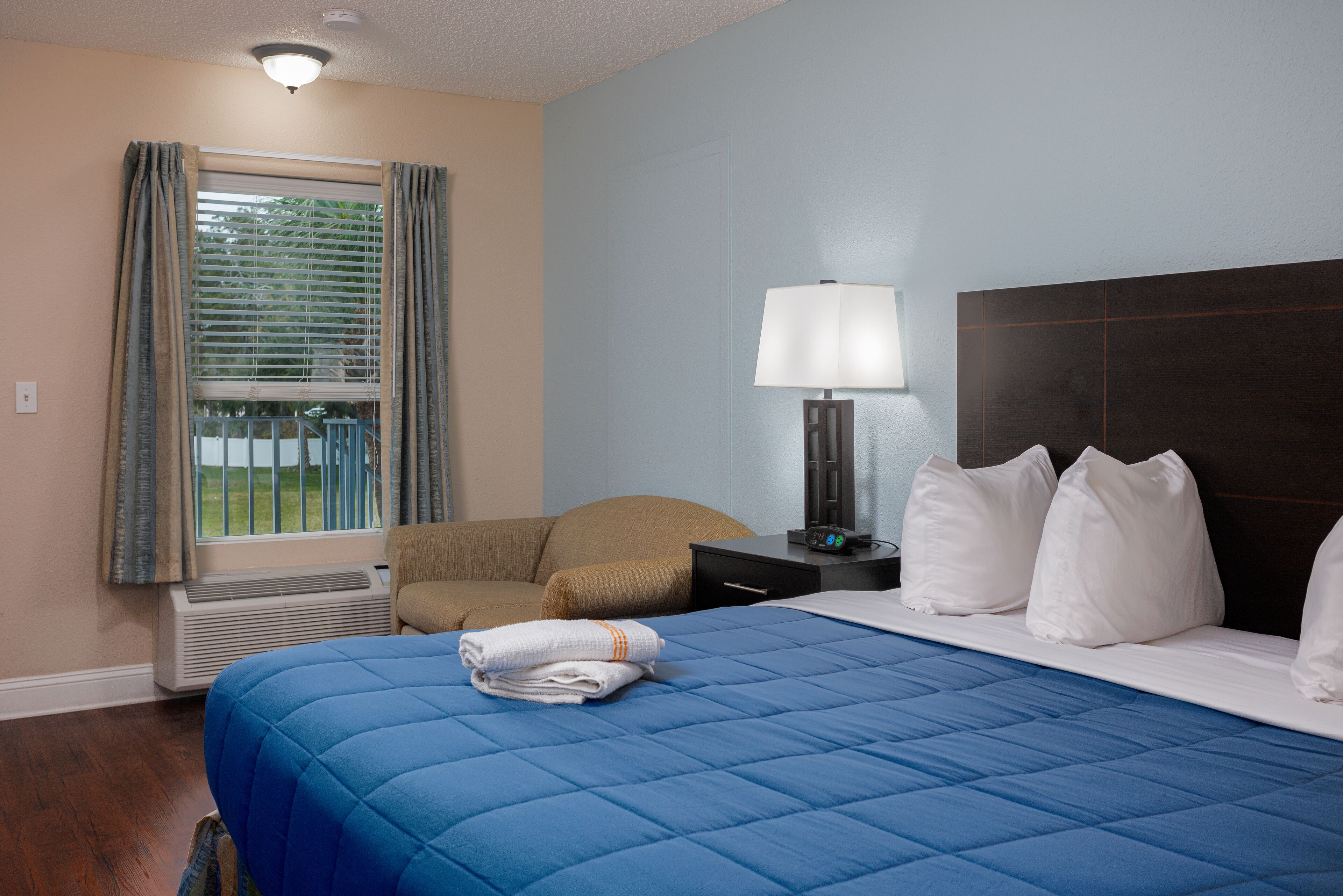 Habitación de hotel con cama, una silla, mesita de noche, lámpara y ventana con cortinas.