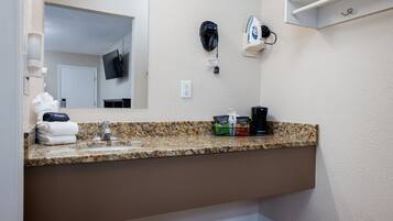 A bathroom with a granite countertop, a sink, a mirror, a towel rack, a coffee maker, and a wall-mounted hairdryer.