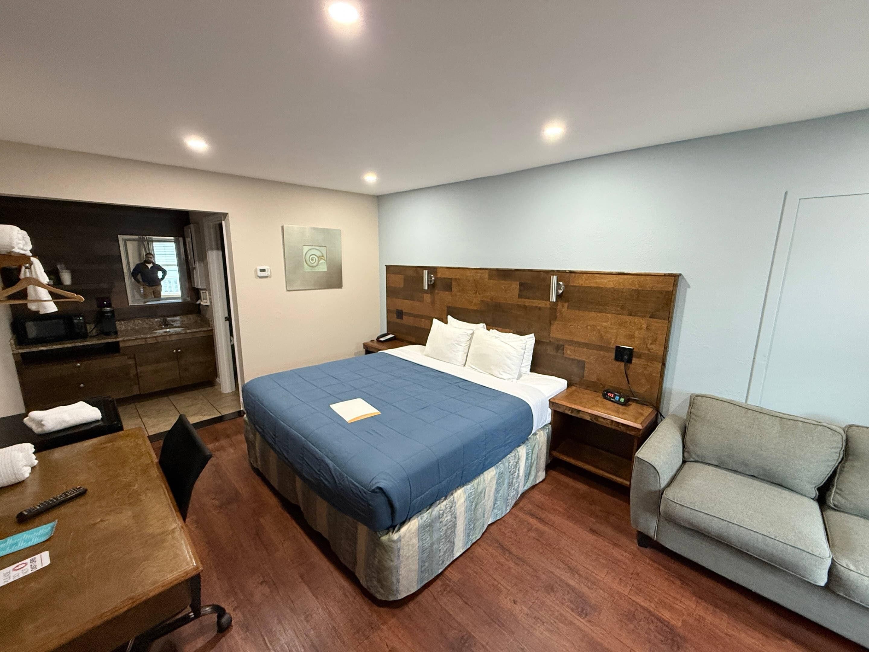 A hotel room with a large bed, a wooden headboard, a gray armchair, a desk with a computer, and a kitchenette area.