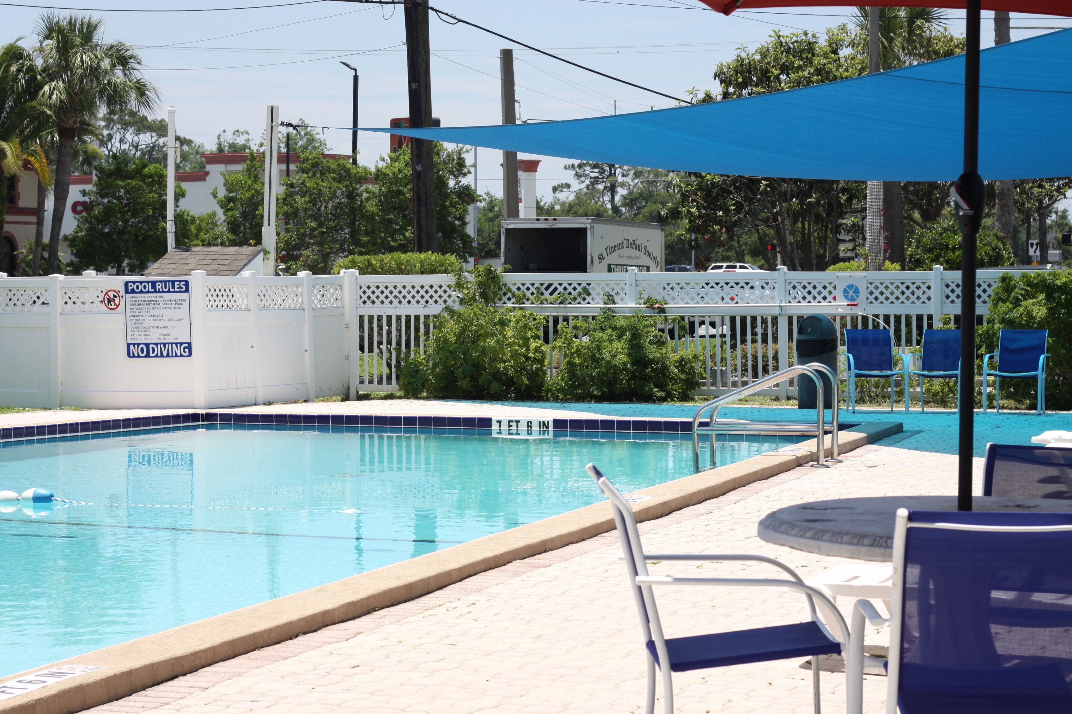 Una piscina con valla blanca, sillas de descanso y un toldo azul.