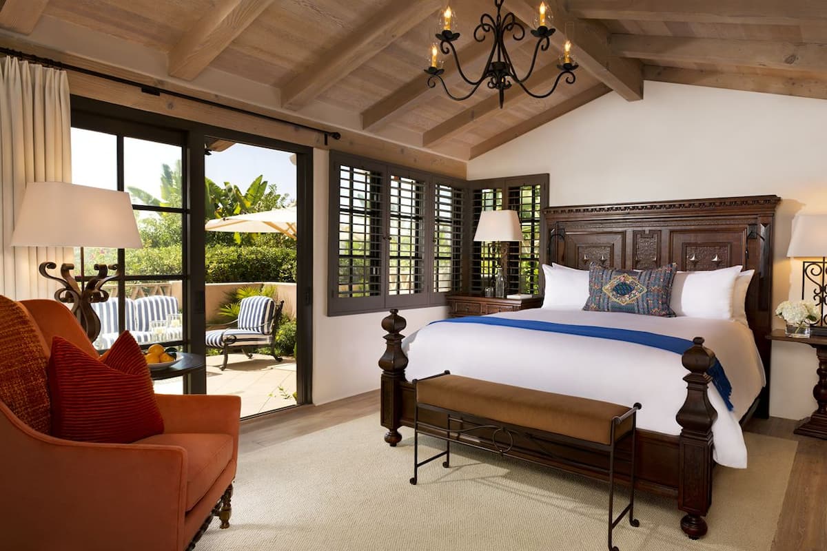 A spacious bedroom with a large bed, a bench, a chair, and a view of the outdoors through large windows.