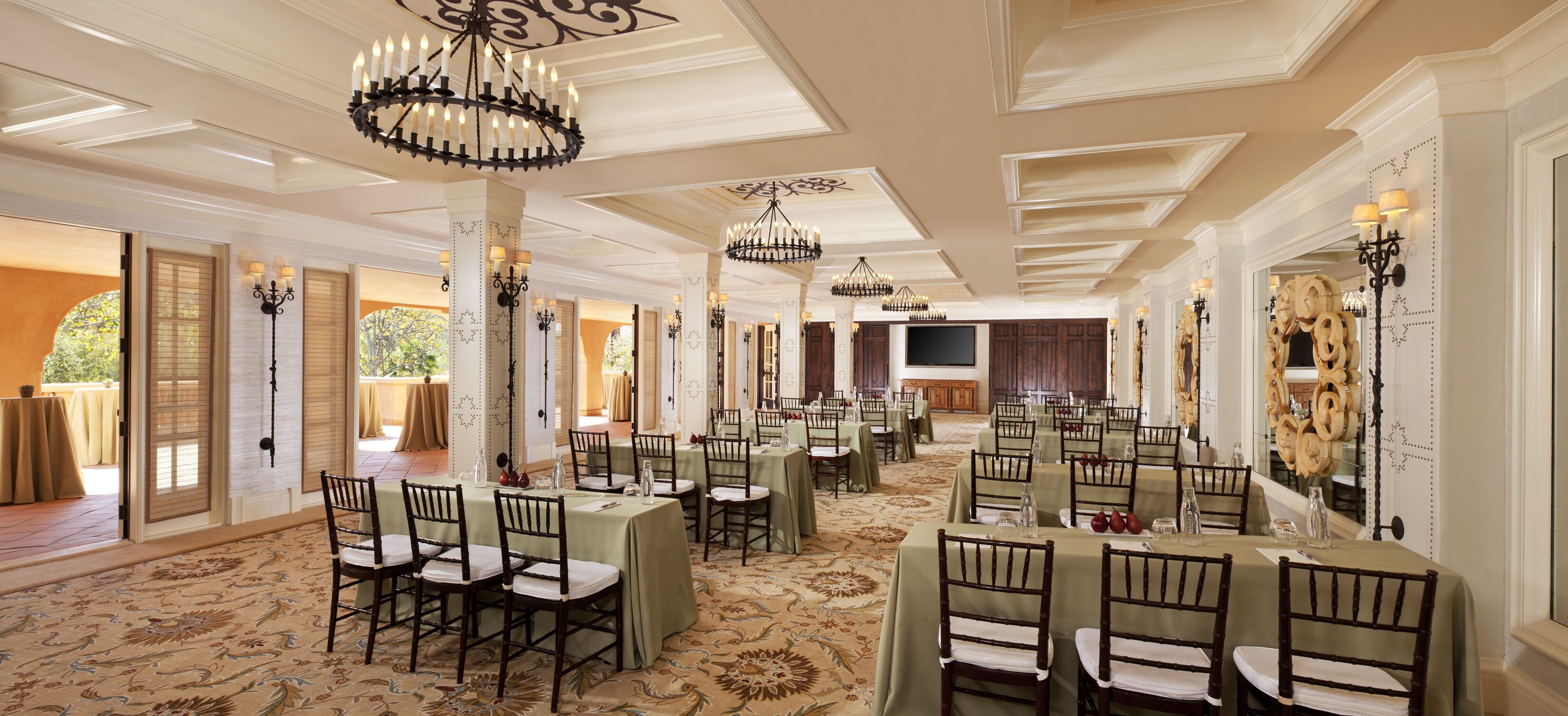 A spacious banquet hall with multiple tables and chairs, elegant chandeliers, and a large television screen.