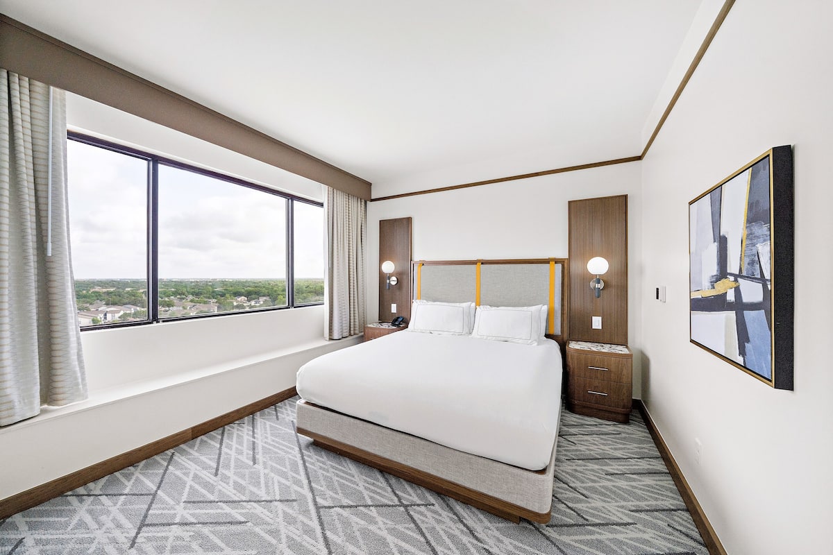 A hotel room with a large bed, a view of the city, a wooden headboard, and a nightstand with a lamp.