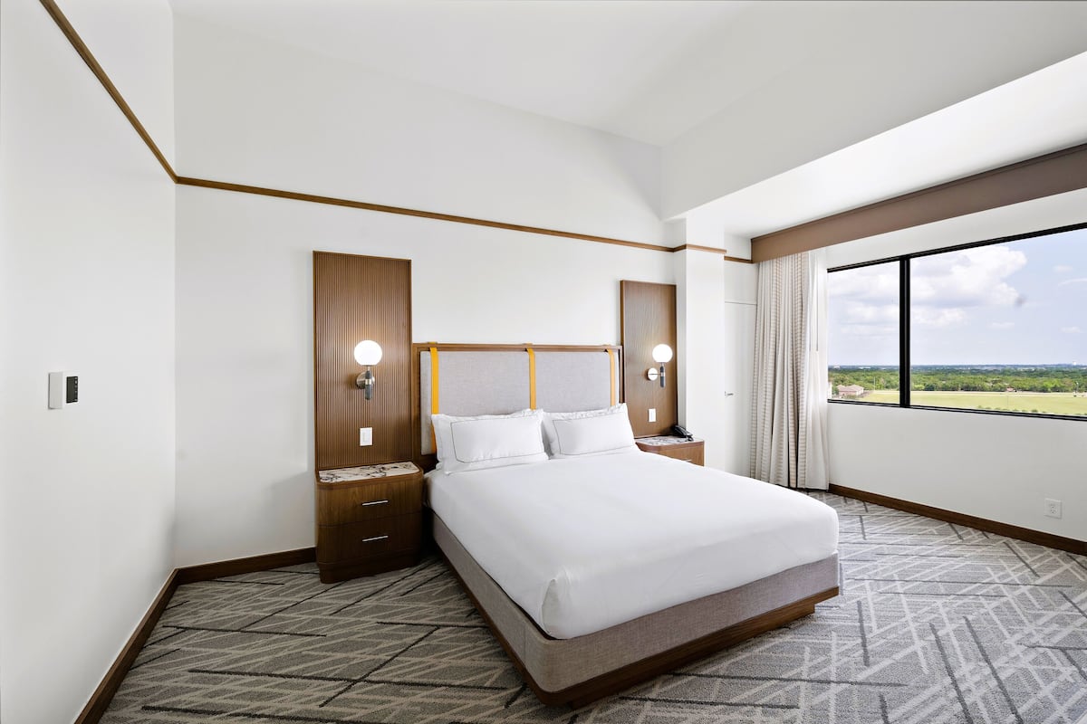 A hotel room with a large bed, wooden headboard, two bedside tables with lamps, a large window with a view of the outdoors, and a carpeted floor.