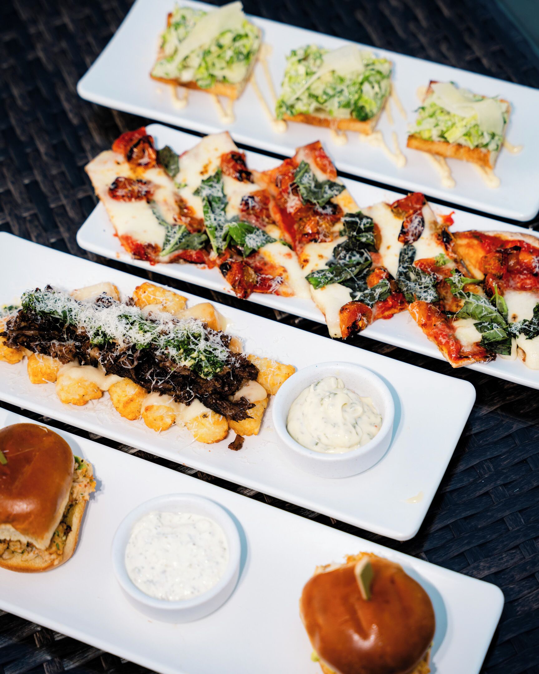 A variety of dishes on white rectangular plates, including pizza and a burger, with small bowls of dipping sauces.