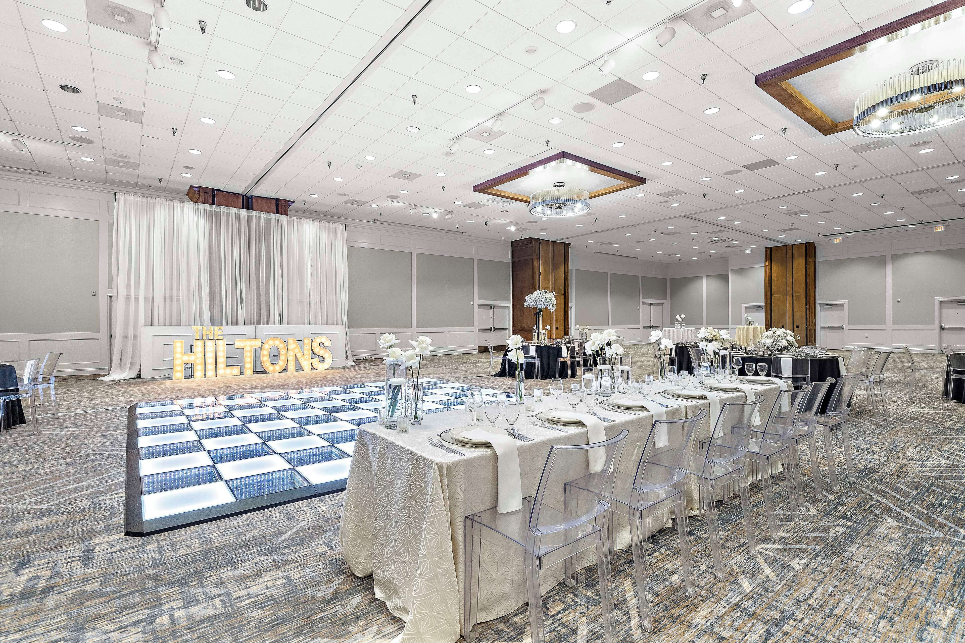 Un amplio salón de banquetes con largas mesas dispuestas para eventos, una pista de baile y un cartel llamativo de 'Hilton'.