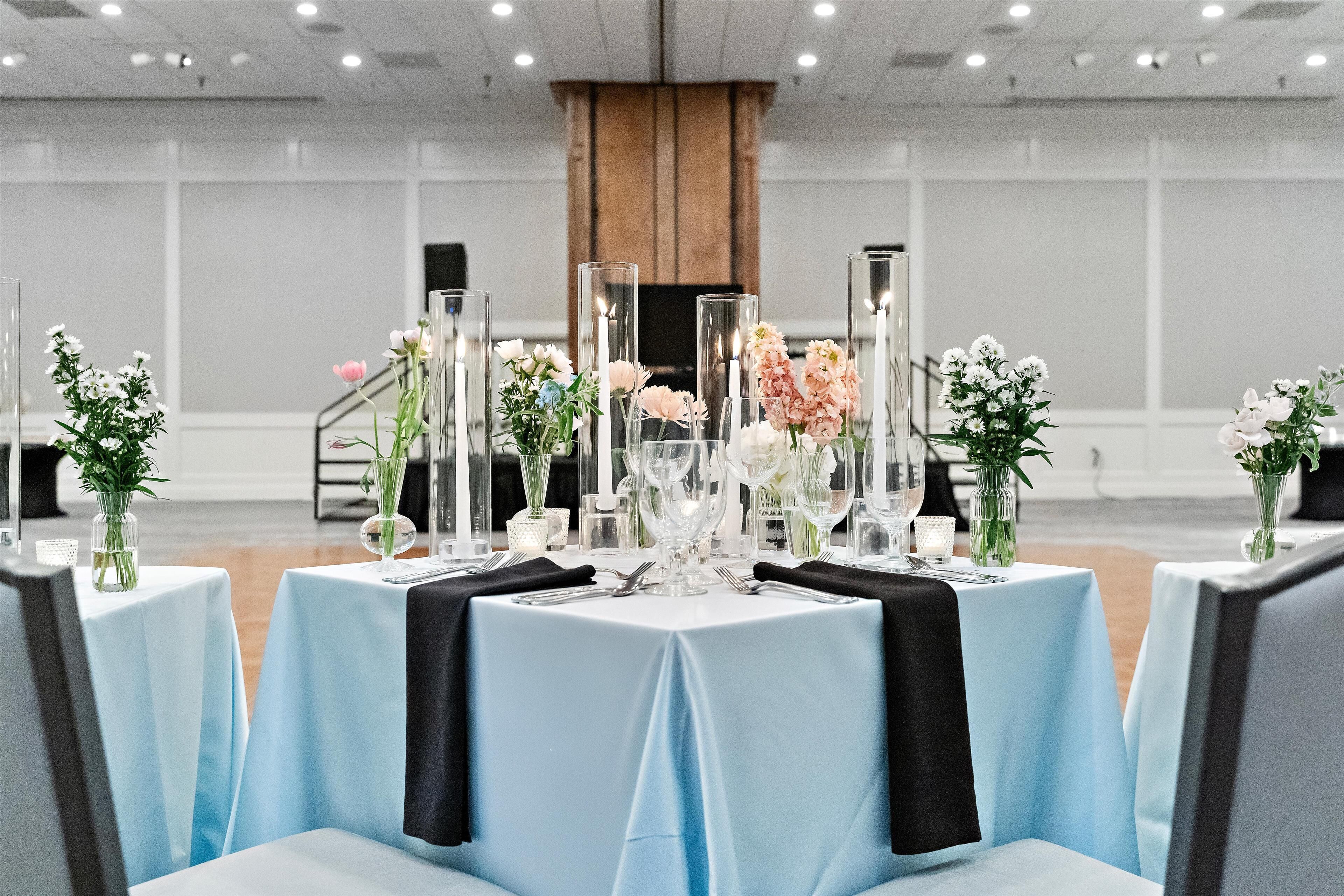 Ein Bankettsaal mit runden Tischen, die mit hellblauen Tischdecken bedeckt sind, jeweils mit Glasware und floralen Mittelstücken bestückt.