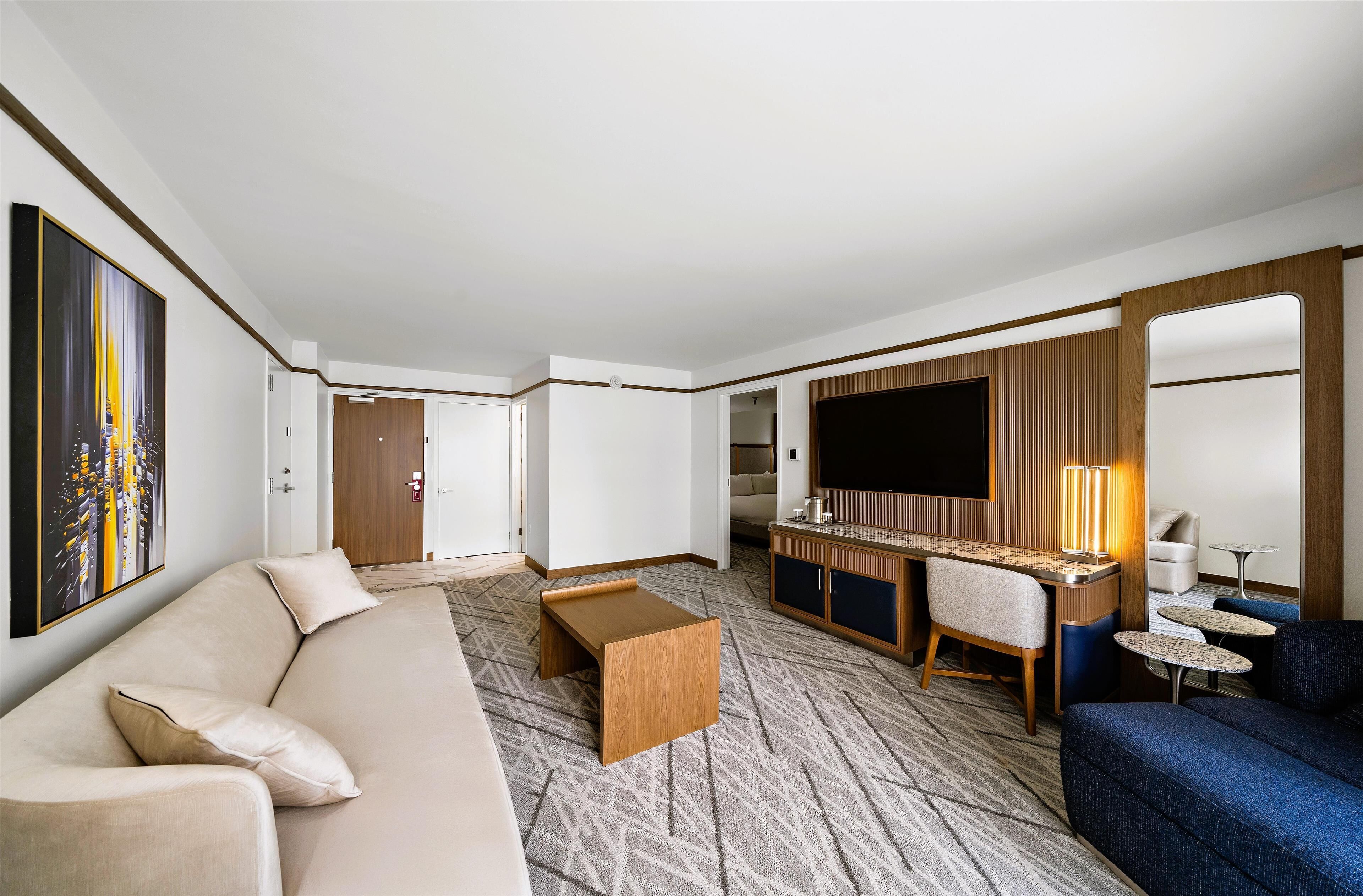 A modern hotel room with a sofa, a small wooden table, a desk with a TV, and a seating area with a lamp.