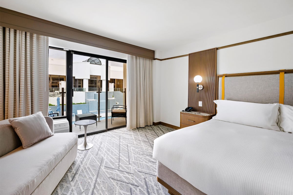 A hotel room with a large bed, a sofa, a small table, a nightstand, and a balcony with curtains.