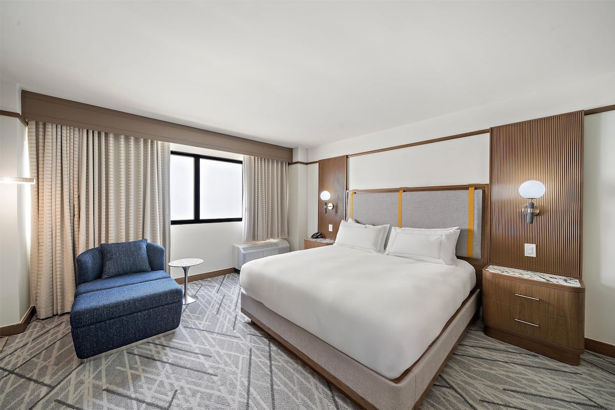 A hotel room with a large bed, a small desk, a chair, and a window with curtains.