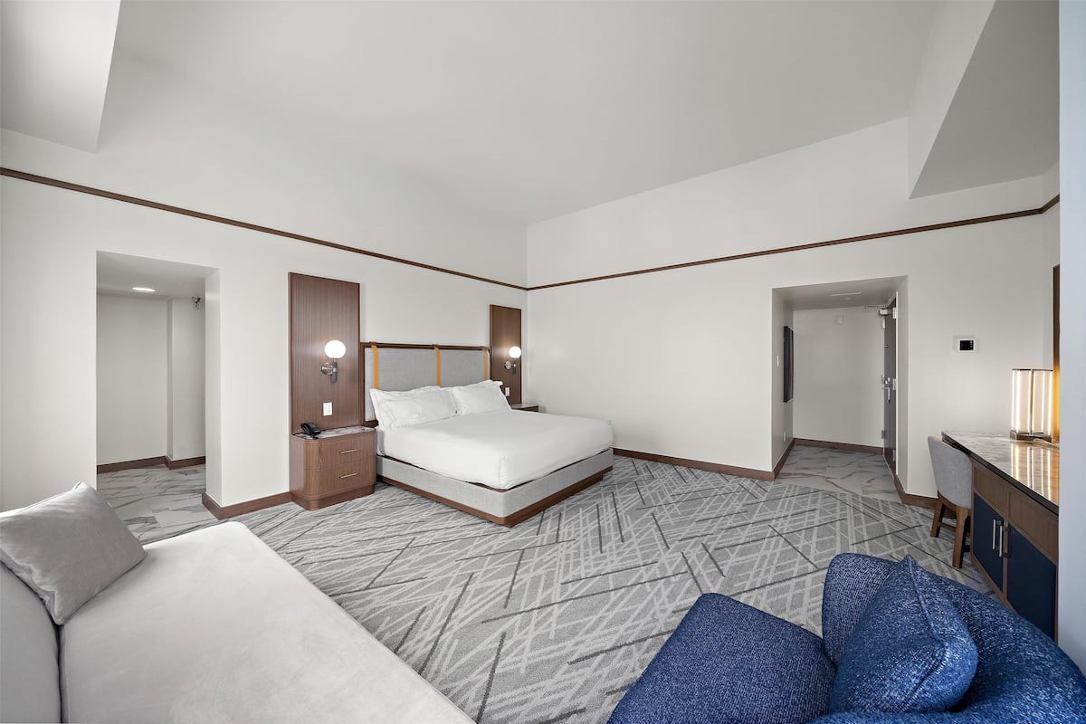 A modern hotel room with a large bed, a sofa, a desk, and a bathroom visible through an open door.