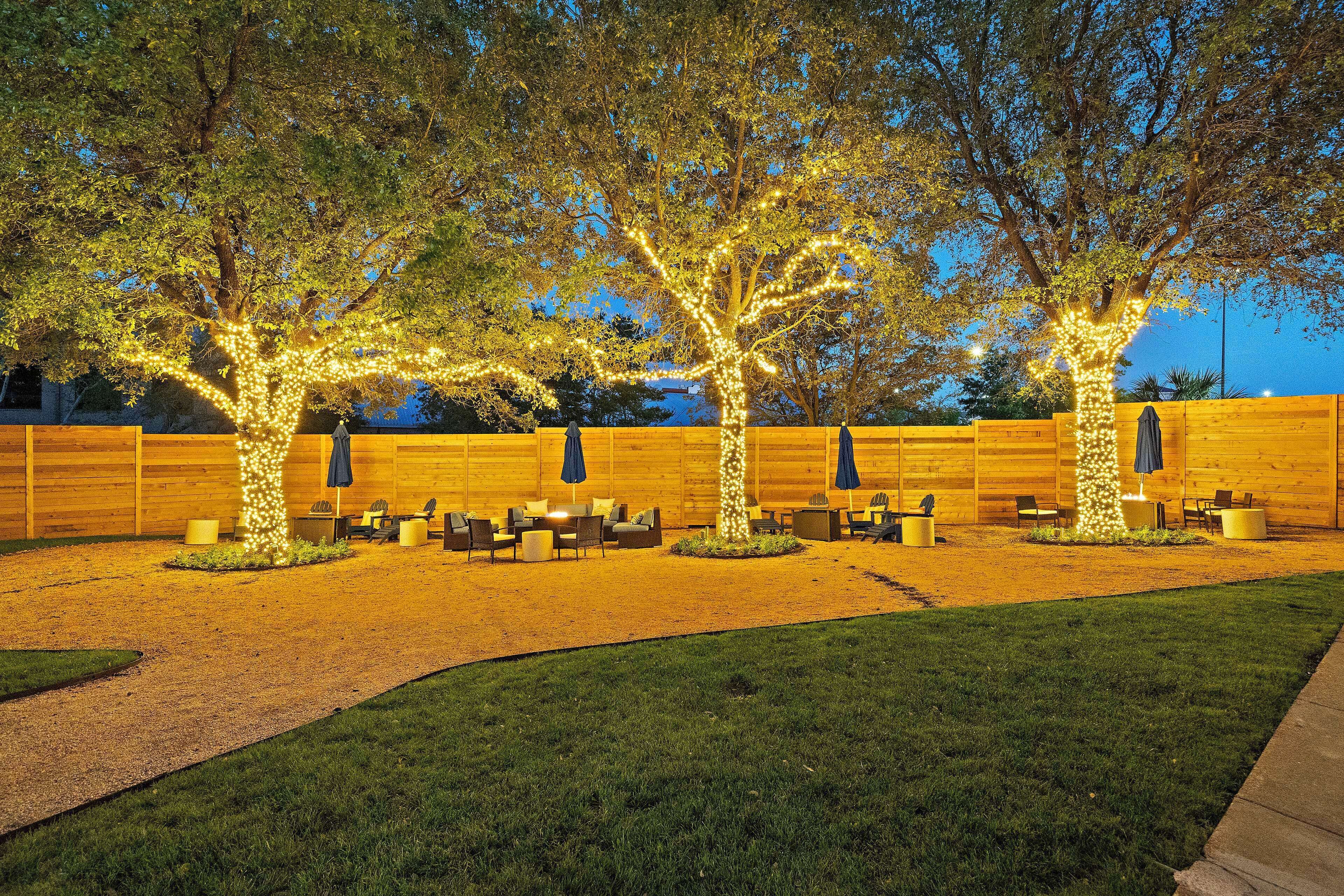 Un área de estar al aire libre con mesas y sillas bajo árboles decorados con luces, una valla de madera y un césped.