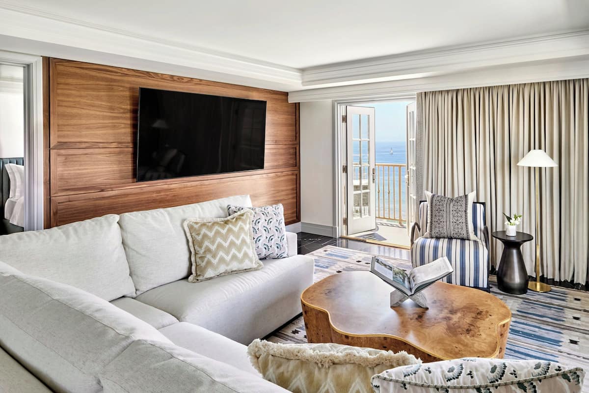 A modern hotel room with a large flat-screen TV mounted on a wooden panel wall, a sofa with patterned cushions, a wooden coffee table with a book, a striped armchair, and a view of the sea through a balcony door.