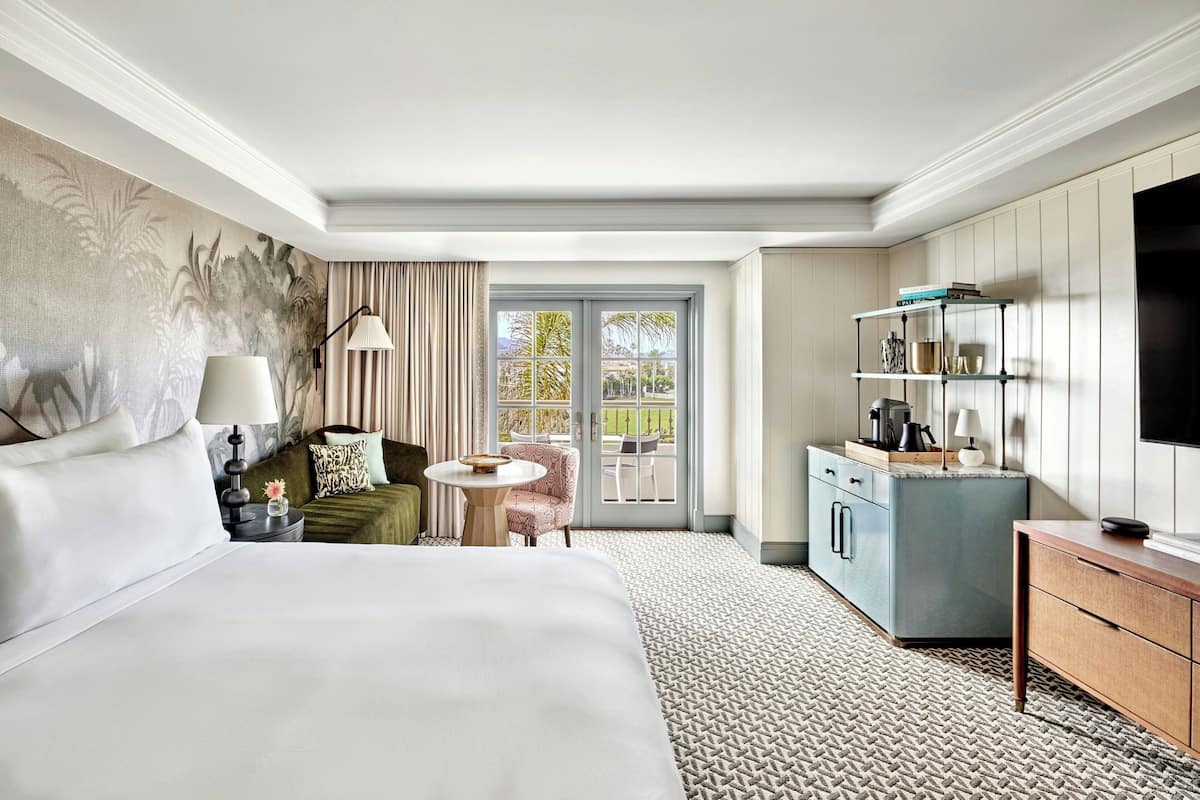 A hotel room with a large bed, a small table with a lamp, a seating area with a chair, a television, and a wardrobe.