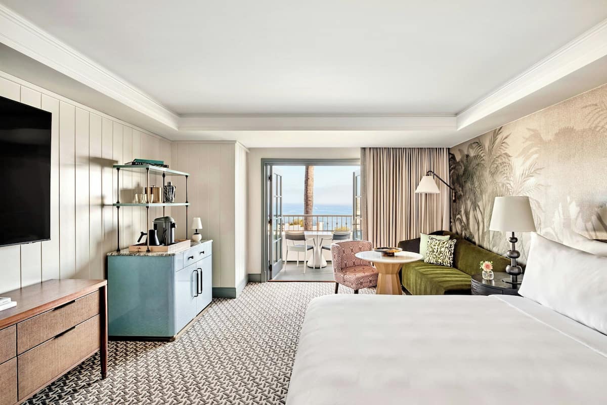 A hotel room with a large bed, a desk with a flat-screen TV, a kitchenette, a balcony with a table and chairs, and a view of the ocean.