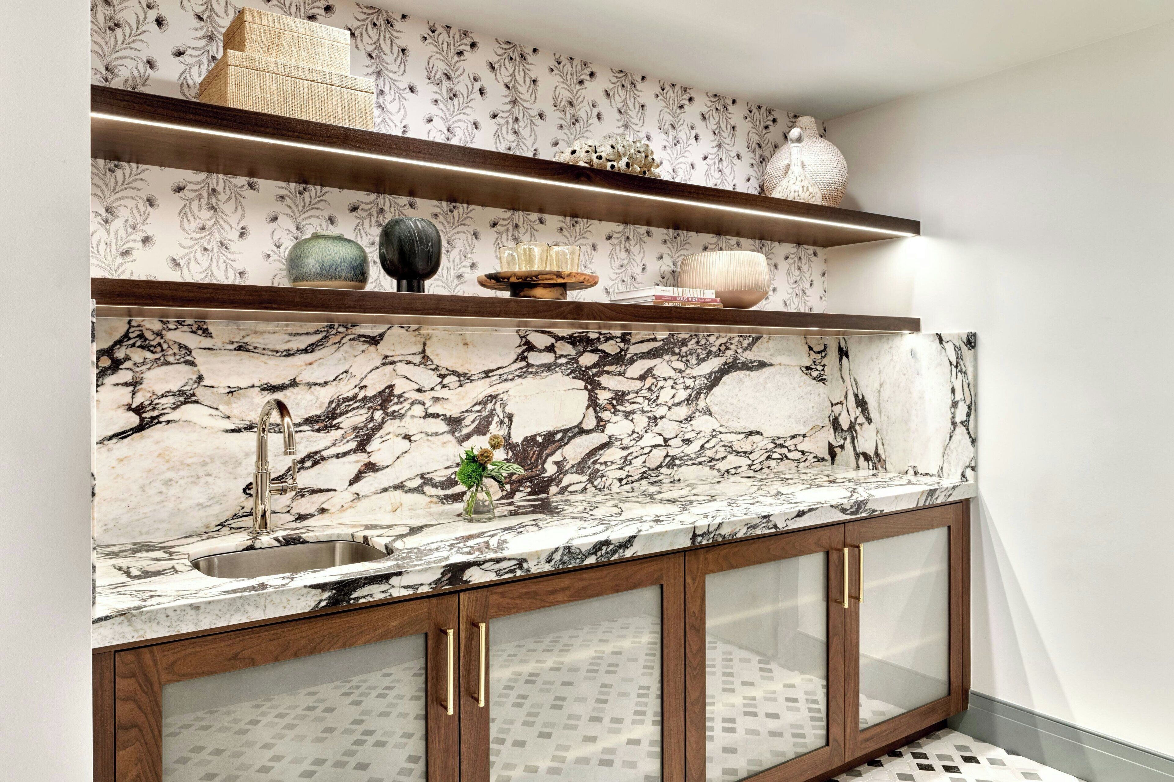A kitchen with marble countertops, a sink, wooden cabinets with glass doors, and decorative items on shelves.