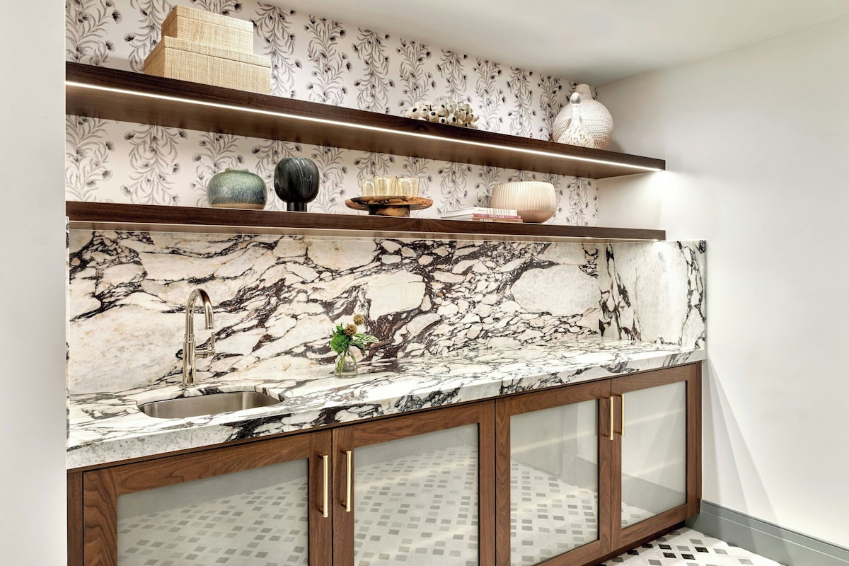 A kitchen with marble countertops, a sink, wooden cabinets with glass doors, and decorative items on shelves.
